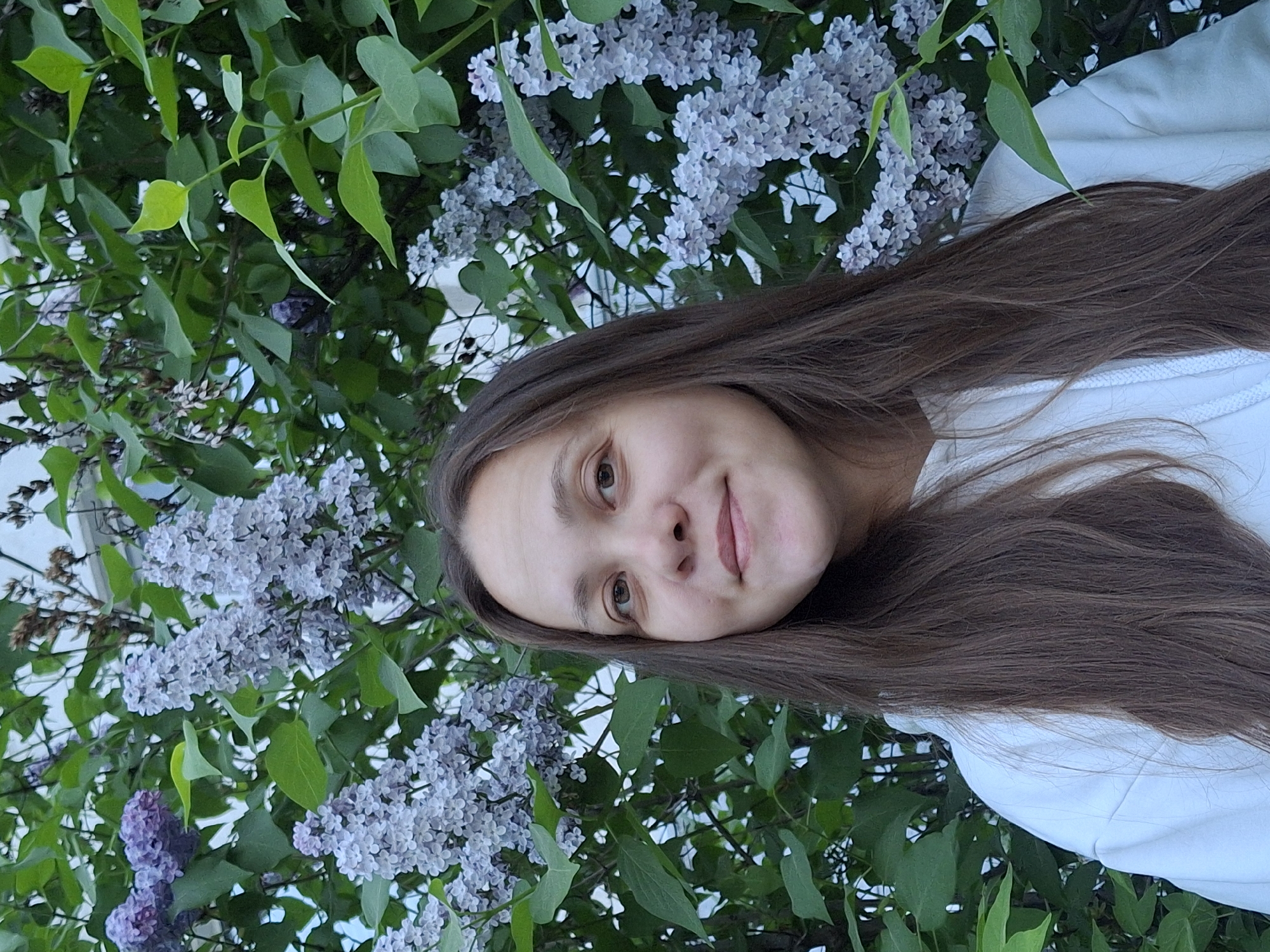 A woman is standing outside next to a lilac bush in Chaikovsky, Russia. The woman has long brown hair, light skin, and is wearing a white top. The lilac bush is full of green leaves and purple and white flowers. The weather appears to be overcast.