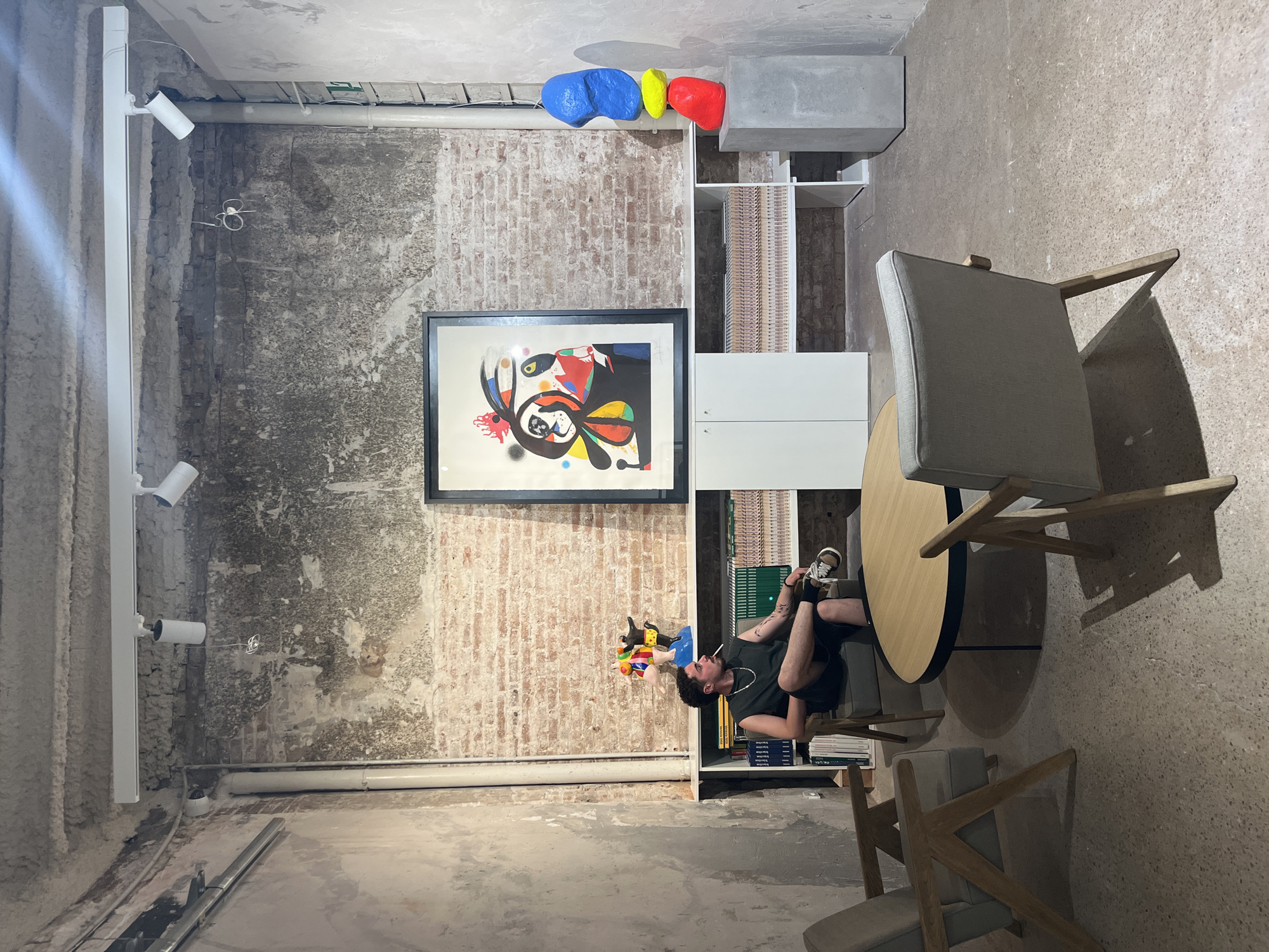 The image depicts a man smoking a cigarette while seated in a chair in what appears to be an art gallery or a creative workspace. The man is wearing a sleeveless dark green shirt and black shorts, with one leg crossed over the other. He has tattoos on his left arm. The room has exposed brick walls and a concrete floor. A round wooden table with black legs is situated in front of him, with two other chairs around it. On a shelf behind the man are several books and a colorful abstract sculpture. To the right, a framed abstract artwork hangs on the wall, and another colorful stacked sculpture is displayed on a concrete pedestal. The overall aesthetic is industrial and modern. While no specific location in Barcelona is provided, the style of the space, with its blend of raw industrial elements and curated art, is consistent with the city's vibrant contemporary art scene.