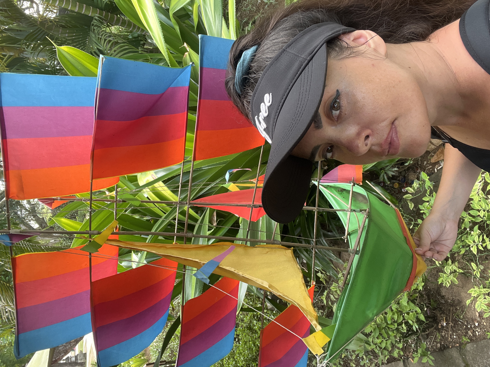 A woman wearing a black visor with white text reads "Free" is looking down at the camera. She has long dark hair, dark eyeliner, and a piercing in her nose. She is wearing a black tank top. In front of her are several colorful kites, possibly arranged like sails on a boat. The kites have horizontal stripes in shades of blue, purple, red, orange, and yellow. Some of the kites are green. The kites are attached to a metal frame. The background consists of lush green foliage and some trees, suggesting an outdoor setting, likely in a tropical location. The scene appears to be taking place during the daytime, with natural light filtering through the leaves. The woman's expression is relaxed and perhaps slightly curious. The overall mood is casual and summery. The location context provided is Kuta Selatan, Indonesia.