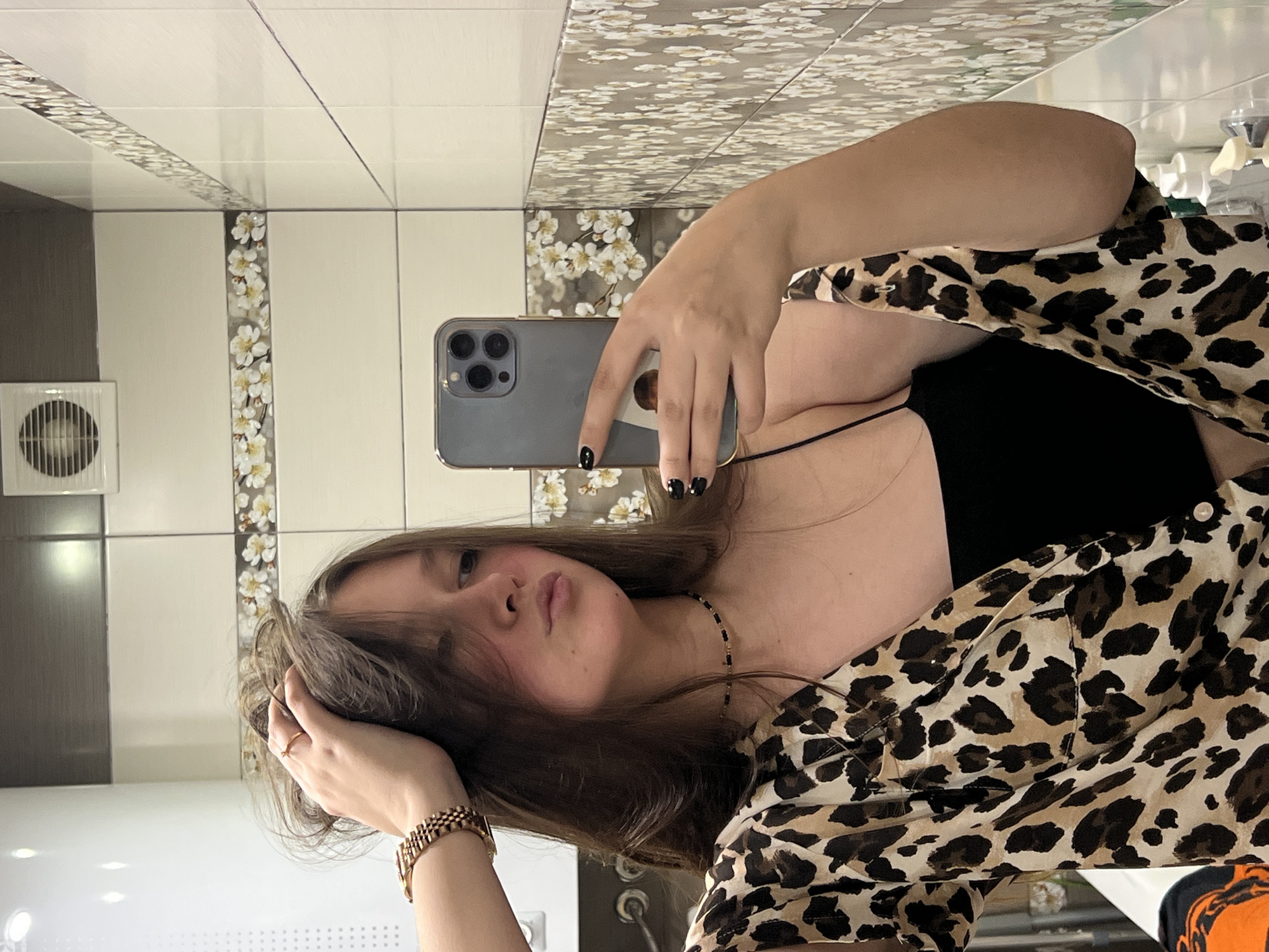 A woman is taking a selfie in what appears to be a bathroom. She is holding a gray smartphone with three visible camera lenses. The woman is wearing a leopard print shirt and a black top underneath. She has long brown hair and is wearing a gold bracelet and necklace. The background features tiled walls with decorative floral patterns. There is also a ventilation fan on the wall. The location is Chaikovsky, Russia.