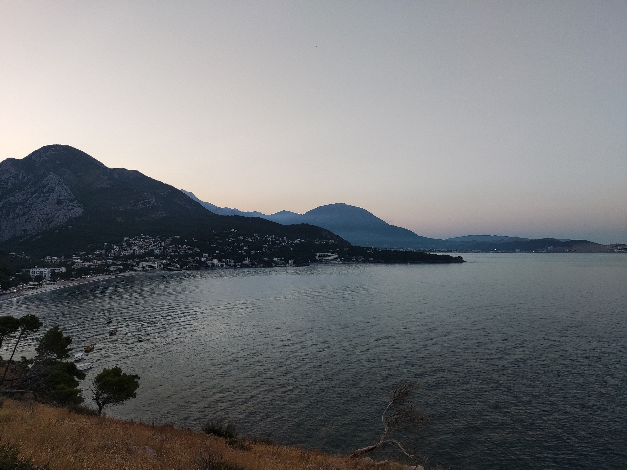 The image depicts a serene coastal scene at dusk, overlooking the city of Sutomore, Montenegro. In the foreground, dry, golden grass covers a sloping hillside. Several sparse, dark-leafed trees are scattered across the hill, some with branches reaching towards the sea. The middle ground is dominated by the calm, rippling surface of the Adriatic Sea, its color a deep grey-blue. A few small boats are anchored or drifting on the water. Along the shoreline, a white sand beach is visible, lined with buildings and the darker green foliage of trees. Further inland, the hillside is densely populated with buildings, forming a town that stretches along the coast. In the background, large, dark mountains rise, their peaks silhouetted against a pale, twilight sky that transitions from a light grey at the horizon to a lighter shade at the top of the frame. The overall impression is one of tranquility and natural beauty, with the human settlement integrated into the landscape.