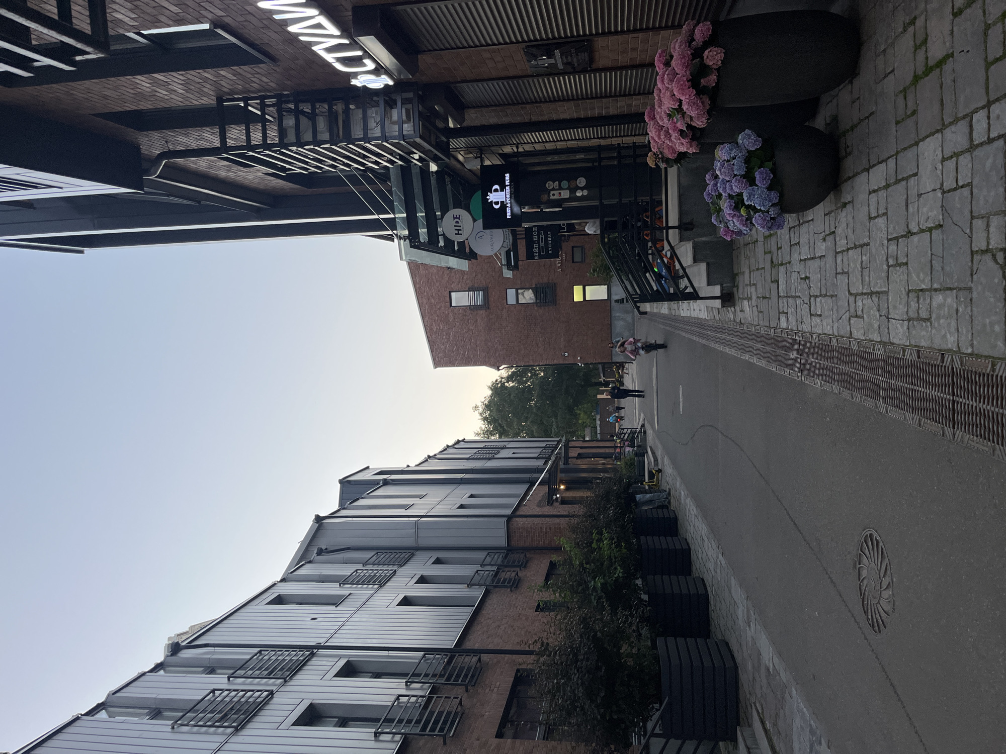 The image depicts a pedestrian alleyway in Khimki, Russia, lined with modern brick and metal buildings. On the left, a multi-story building features dark metal siding with rows of windows and small balconies. Planters with dark green foliage are placed at the base of this building. The right side of the alley is dominated by a brick building with a glass awning leading to an entrance with several signs, including "HİDE," "AURAMED," "BEЙN-ШОП," and "FRET & PORTER FURS." Neon signage with Cyrillic letters is visible on this building, reading "СТУДИ" which likely means "STUDIO." Potted hydrangeas in shades of pink and blue are displayed in large dark planters at the entrance. The alley pavement is a mixture of asphalt and cobblestones, with metal grates for drainage. In the background, a few individuals are walking, one on a bicycle, and a yellow electric scooter is parked near the entrance of the brick building. The sky is a clear, pale blue, suggesting it is either early morning or late evening.