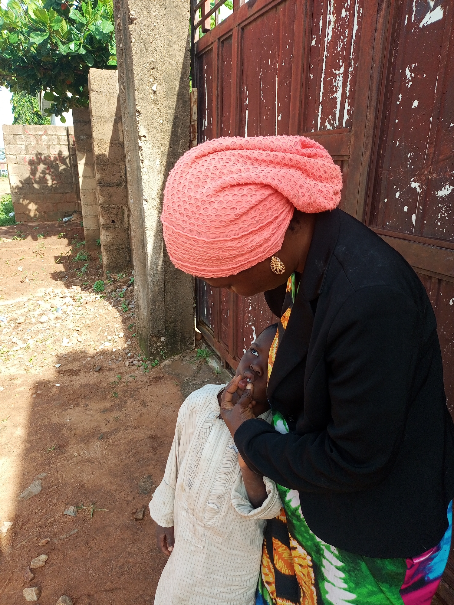 In Tempure, Nigeria, a person wearing a bright pink headscarf and a black jacket is tending to a child. The child, dressed in a traditional light-colored garment, is looking upwards as the person gently holds their mouth. A vibrant, patterned fabric is visible around the person's shoulders, suggesting it's part of their attire. The background features a weathered, reddish-brown metal gate, a concrete pillar, and a brick wall, indicative of an outdoor, possibly residential or communal area. The ground is a mix of reddish-brown earth, small stones, and sparse vegetation, suggesting a semi-arid or well-trodden environment.