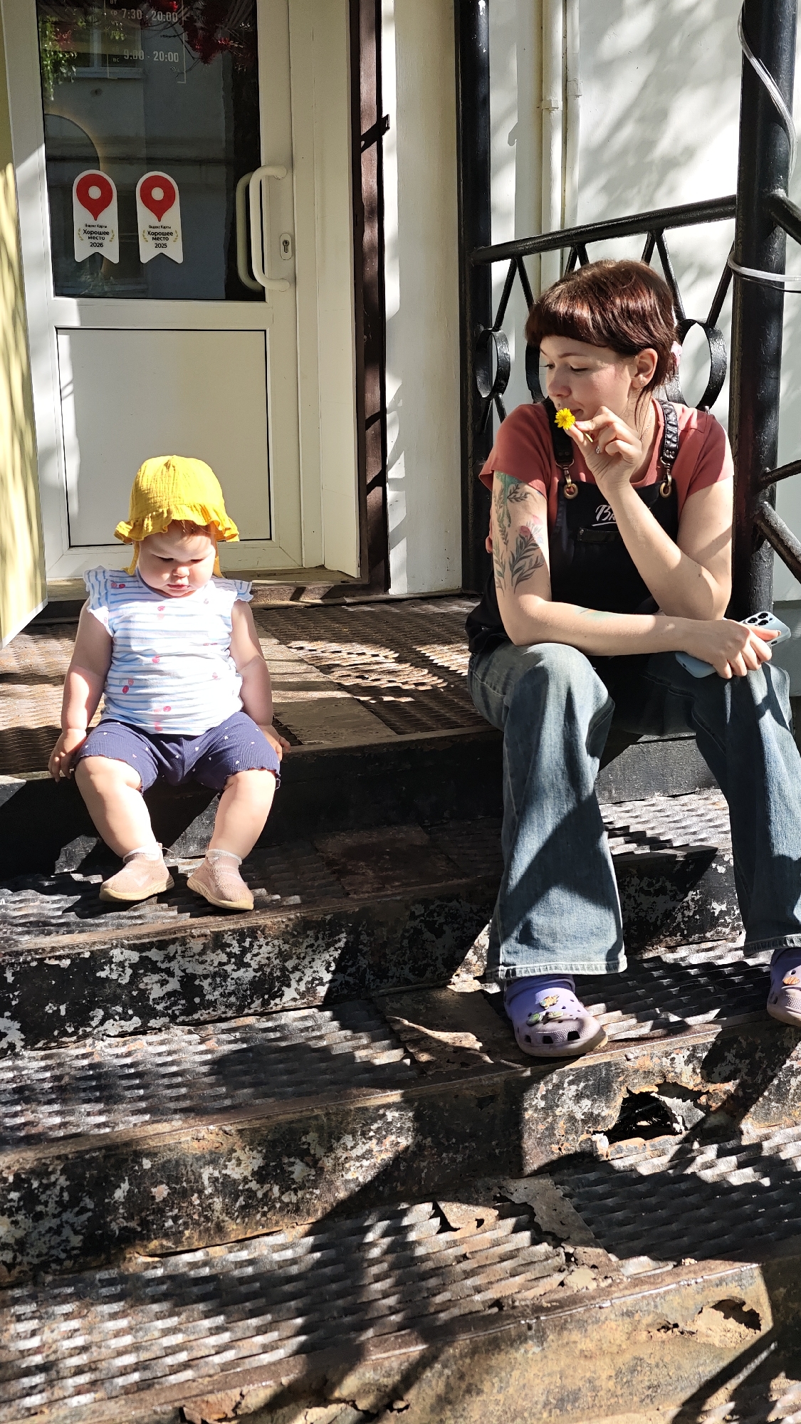 A woman and a baby sit on the metal steps outside a building with a glass door marked with signs in Russian. The signs read: “Super-card” “Good place” 2026 and 2025. Hours listed: 7:30-20:00. 9:00-20:00. The baby sits alone on the first step, facing forward, wearing a yellow sun hat, patterned shirt and matching blue bottoms. The woman sits with one leg on the higher step, and one leg on the lower step with a partially visible hole in the corroded metal. She is holding a yellow flower up to her face, with her other hand holding a cellphone. She wears blue jeans, purple shoes, a pink t-shirt, black apron and has a tattoo on her arm. The location is Chaikovsky, Russia.