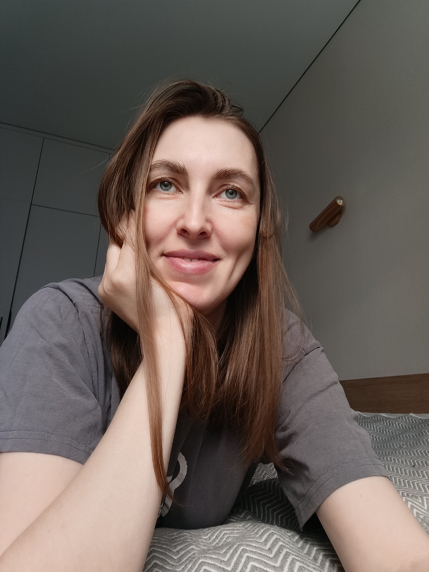 A medium close-up shot depicts a fair-skinned woman with shoulder-length brown hair and blue eyes. She is propped on her left arm, resting her cheek on her hand, and smiling slightly. She is wearing a gray t-shirt. The background suggests an interior setting with a gray wall and a wardrobe cabinet visible on the left. A small, cylindrical wooden wall lamp is mounted on the wall. The textured bedding beneath her arm indicates she is likely lying on a bed. While the image provides an interior scene, there is no specific information in the image to confirm the location as the city of Chaikovsky, Russia.