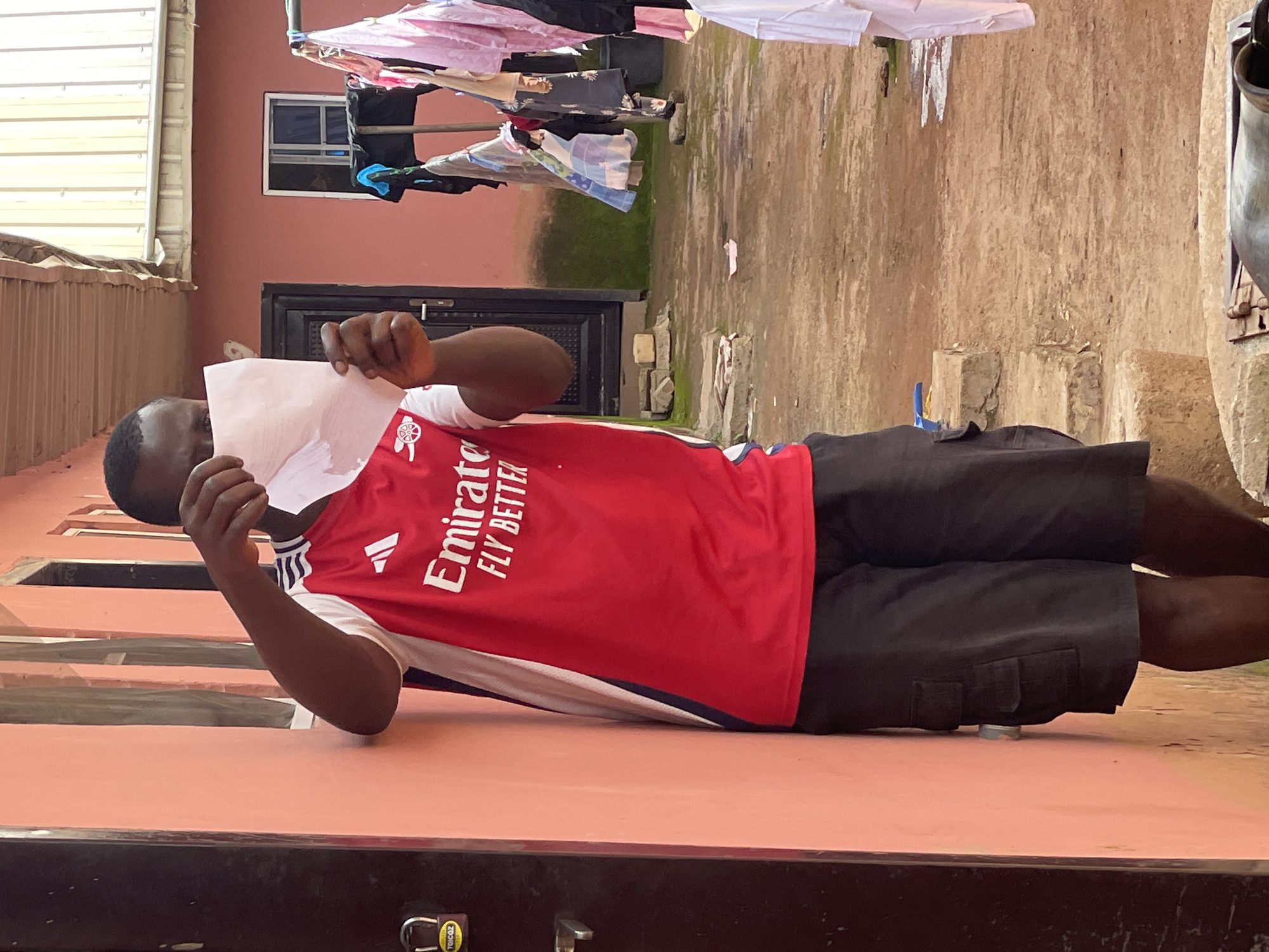 A Black man, wearing a red Arsenal soccer jersey and black shorts, is lying on his side. He is holding a white piece of paper up to his face, obscuring his mouth and nose. The man is in a dimly lit outdoor area with a reddish-brown wall behind him. Laundry is hanging on a clothesline above and behind him. In the background, there appears to be a doorway with a black door and a small window. The ground is dirt and has some patches of grass. The scene appears to be a residential area in Durimi, Nigeria. The time of day is unclear, but it could be daytime. The weather appears to be dry. The man's emotions are not discernible due to the paper covering his face. The visible text on the jersey reads "Emirates FLY BETTER". There is also a small yellow tag with illegible text on a lock in the lower foreground.