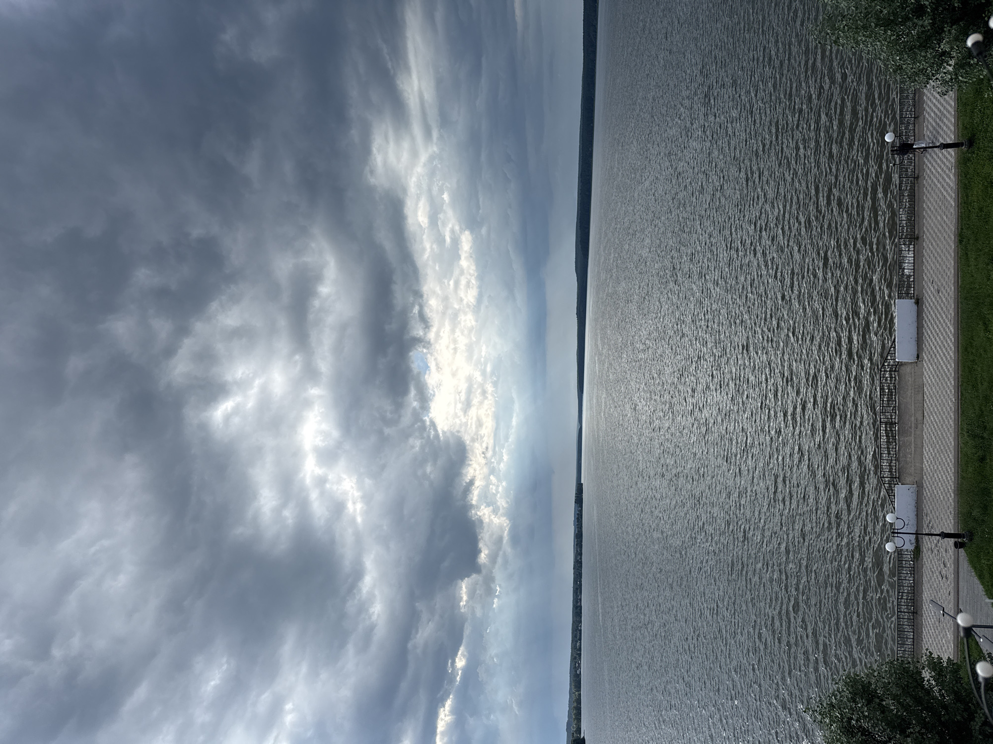 The image depicts a wide, calm body of water, likely a reservoir or a large river, under a heavily overcast sky. Dramatic, dark clouds dominate the upper portion of the frame, with shafts of light breaking through in places, suggesting an approaching storm or recent rain. The water's surface is rippled, indicating a gentle breeze. In the distance, a tree-lined shore stretches across the horizon, with a faint hint of buildings or settlements on the left side.
In the foreground, a paved walkway runs along the edge of the water. A decorative black metal fence separates the walkway from the water. Several lampposts with white spherical lights are visible along the walkway, along with white benches. Green grass borders the paved area. The scene is devoid of people.
The location is identified as Votkinsk, Russia, and the body of water is the Votkinsk Reservoir, formed by the damming of the Kama River. The viewpoint appears to be from an elevated position, looking down towards the reservoir and the embankment. The overall atmosphere is somewhat moody and dramatic due to the weather conditions.