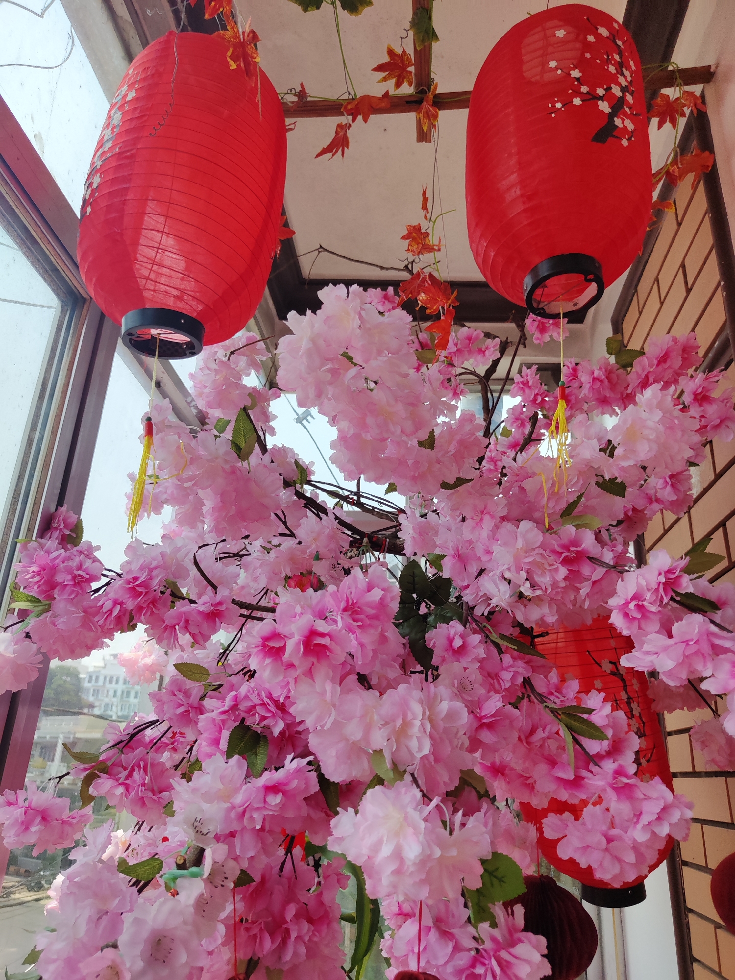 The image shows an indoor scene in Da Lat, Vietnam, decorated with red lanterns and pink cherry blossoms. Two large, red, oval lanterns hang from the ceiling, one plain and one with a white cherry blossom design. Beneath the lanterns, a dense arrangement of artificial pink cherry blossoms fills the frame, obscuring the view of the cityscape outside a window on the left. Orange maple leaf garlands are strung near the ceiling. A brick wall is visible on the right side of the frame. The overall composition suggests a celebratory or festive setting, likely for a holiday or special occasion.