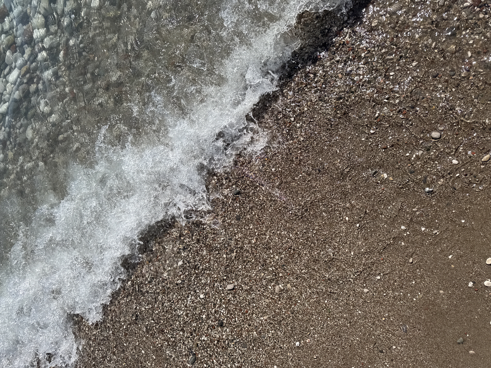 This is a high-angle, close-up photograph of a shoreline at Šušanj, Montenegro. The image captures the dynamic interaction between the water and the beach. In the foreground, a wave is receding from a pebbly shore. The water is clear and shows the texture of the stones beneath its surface, with sunlight glinting off the ripples. The beach itself is composed of small, dark brown pebbles and sand, with occasional larger, lighter-colored stones and some shell fragments visible. A white, foamy edge of the wave laps at the pebbles, creating a frothy texture. The background is dominated by more of the shallow, clear water with visible pebbles on the seabed. There are no people or man-made objects present in the scene. The environment appears to be a natural, coastal setting.