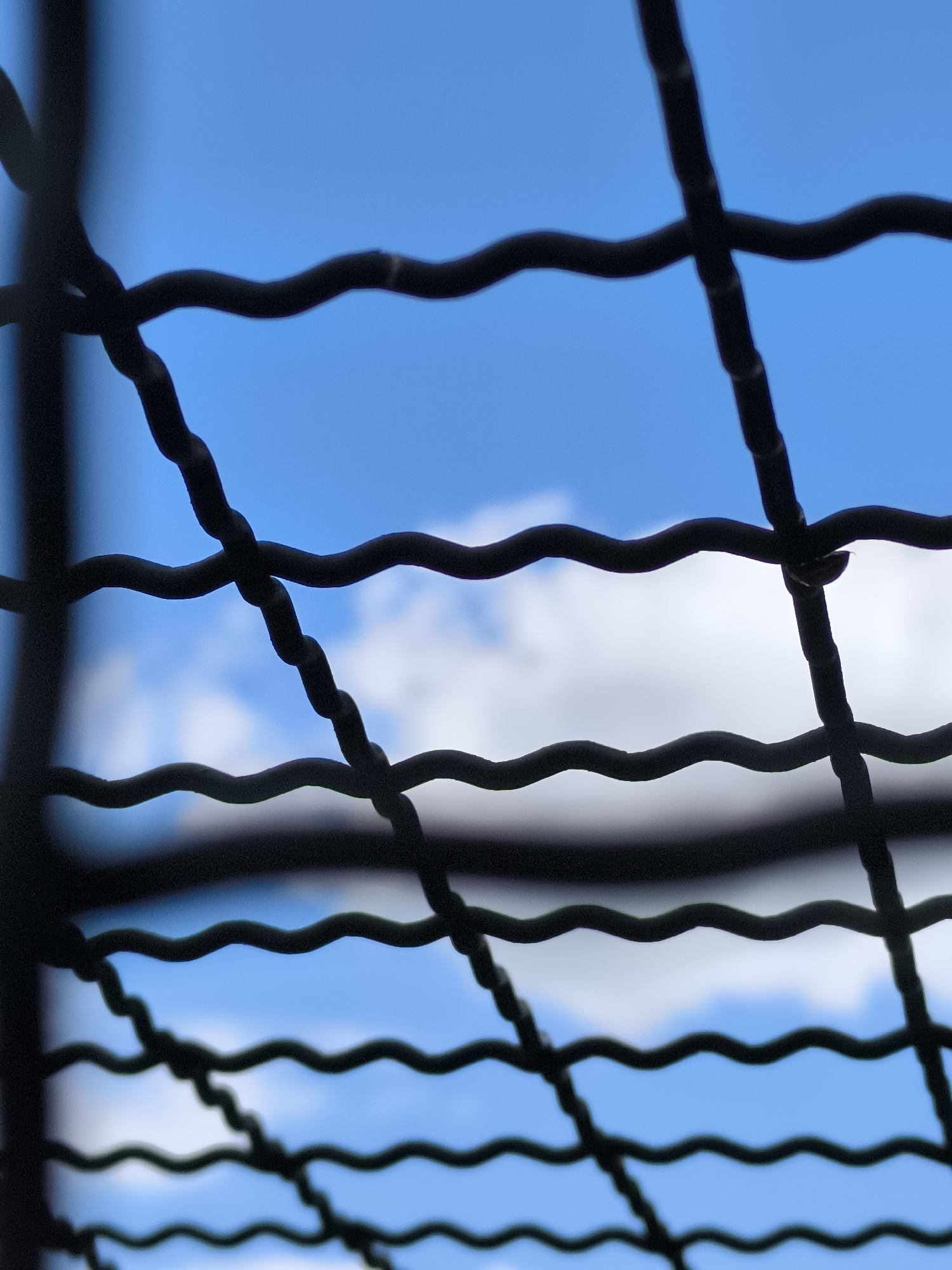 This close-up, abstract photograph captures a section of a dark, metallic grate against a bright blue sky with scattered white clouds. The grate consists of multiple wires, some of which are wavy in shape, creating a grid-like pattern. These wires are in sharp focus in the foreground, obscuring large portions of the background. The sky and clouds are blurred, suggesting a shallow depth of field. The lighting indicates it is daytime, likely sunny due to the clear blue sky and the brightness of the clouds. The scene evokes a sense of looking through a barrier, perhaps a fence or window, towards an open outdoor space. There is no visible text.