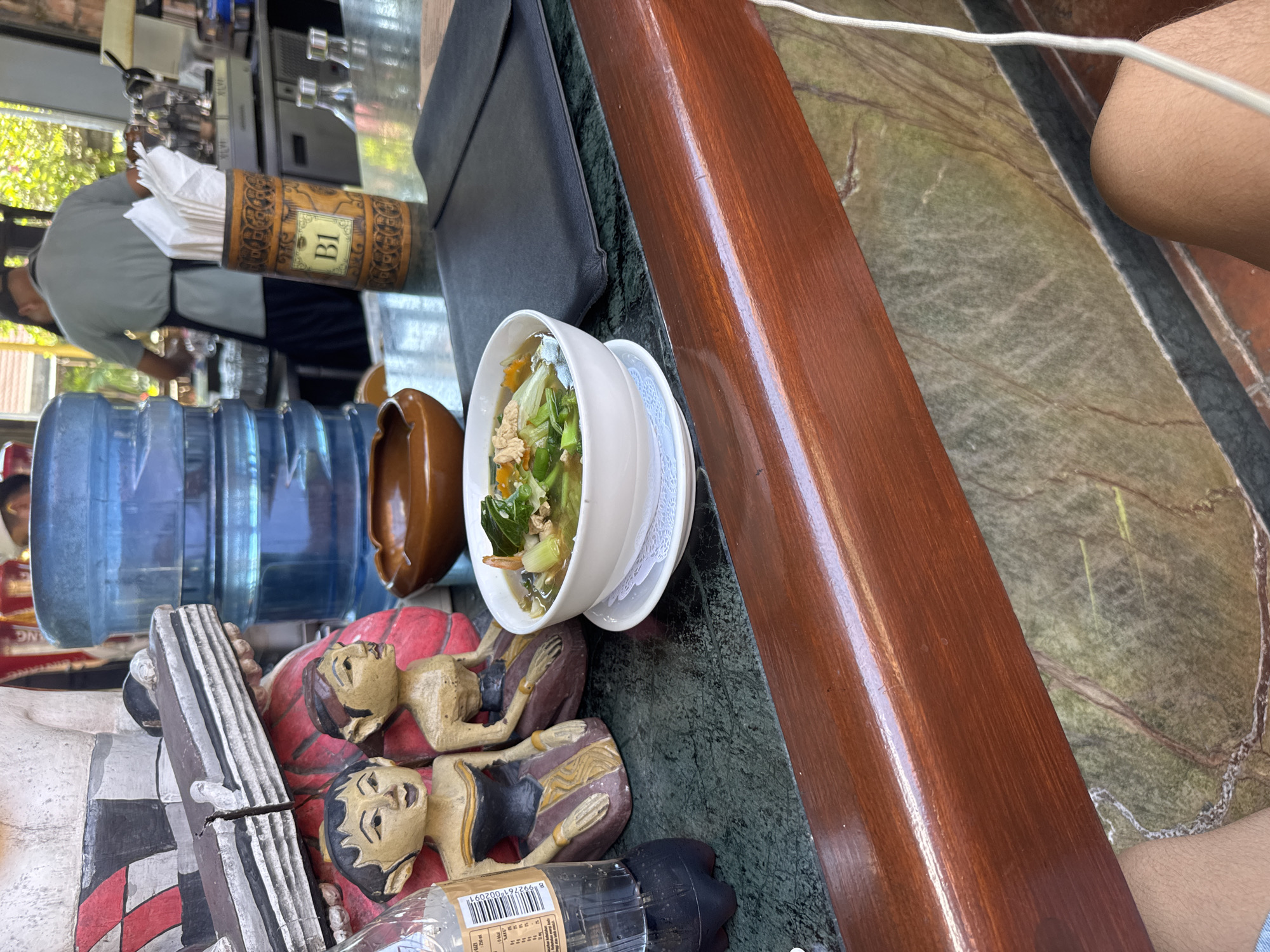 A dining table at a restaurant in Kuta, Indonesia, is partially visible, constructed with a green-marbled surface and a polished wooden edge. A bowl of soup sits on a saucer. Adjacent to the bowl are two carved wooden figurines. A water cooler jug and a brown cylindrical holder for napkins are in the background. A person wearing an apron can be seen behind the table. A menu is resting on the table, partially obscured. A bottle is partially visible below the wooden figurines. The foreground shows part of a decorative item. A person's arm is in the frame, on the right side of the image.
