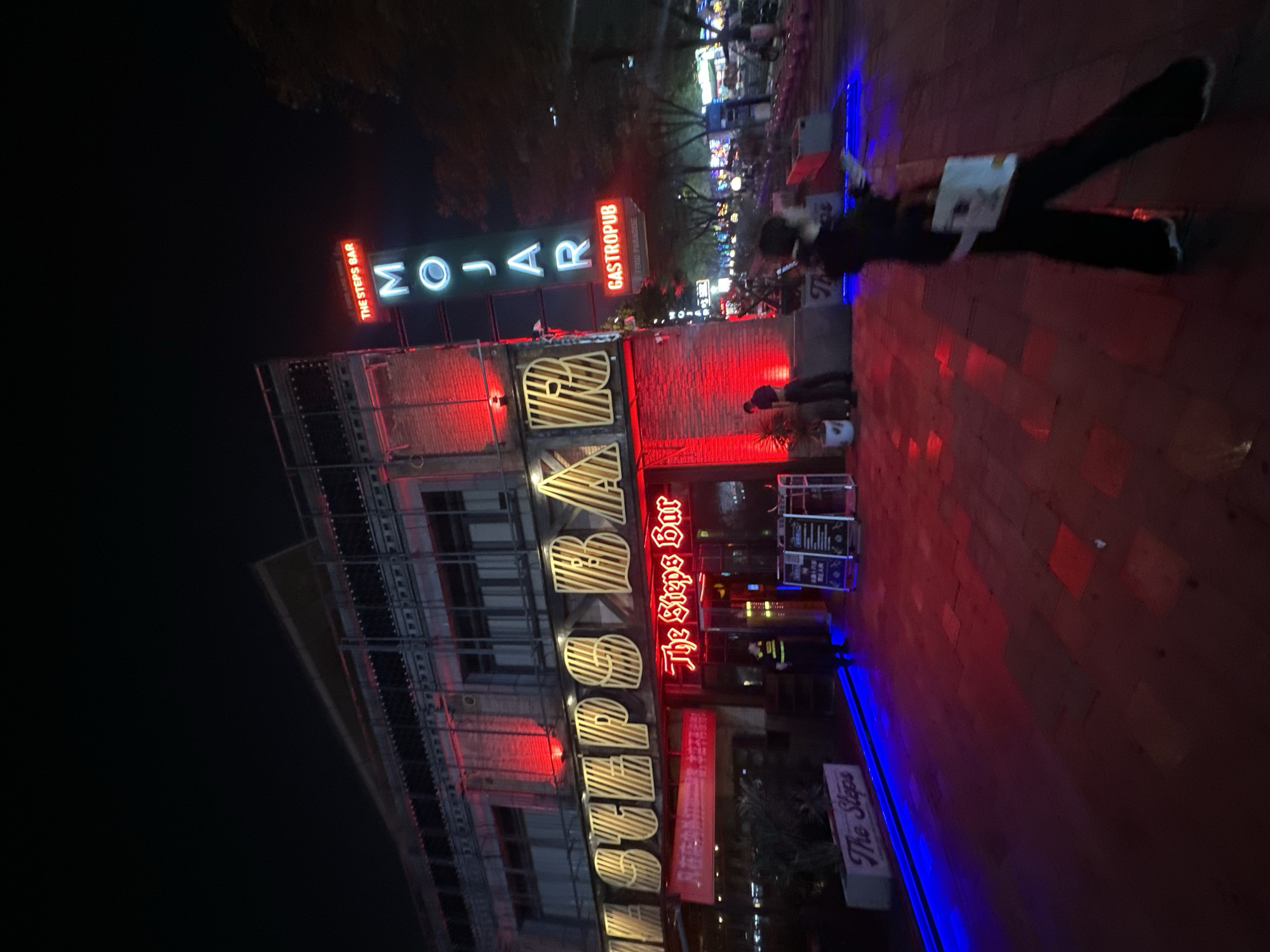 At night, a building with red and blue neon lighting is visible. The building has large, illuminated signs with the words "STEPS BAR" and "MOJAR GASTROPUB". A person in a security uniform stands near the entrance of "The Steps Bar". The ground in front of the building is paved with red bricks. In the background, a street scene with more lights and trees is visible, suggesting an urban environment. A person is walking away from the camera.
