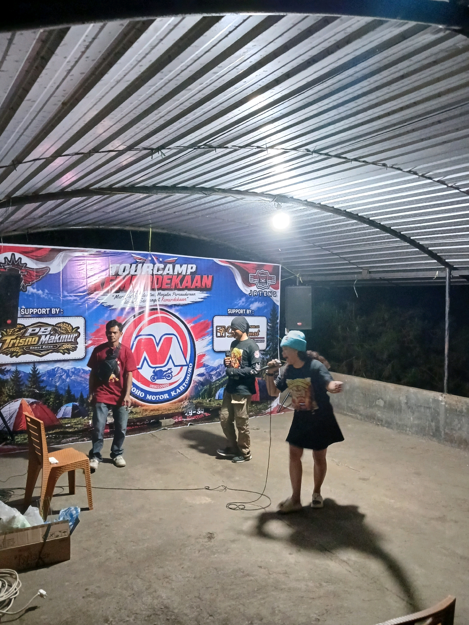 In the city of Berjo, Indonesia, an outdoor event is taking place under a corrugated metal roof. The event appears to be a "Tourcamp Merdekaan," as indicated by a large banner in the background. The banner features a mountainous landscape, tents, and various logos, including one that reads "PB Trisno Makmur" and another with the letter "M" inside a circular emblem with text around it. Three individuals are visible. Two men are standing near the banner, one in a red shirt and jeans, and another in a black shirt and beanie. A third person, wearing a black t-shirt, black skirt, and a light blue beanie, is in the foreground, holding a microphone and seemingly performing or speaking. They are casting a prominent shadow on the concrete ground. A plastic chair is positioned to the left of the frame, and several boxes and coiled cables are on the ground. The overall atmosphere suggests a casual gathering or performance.
