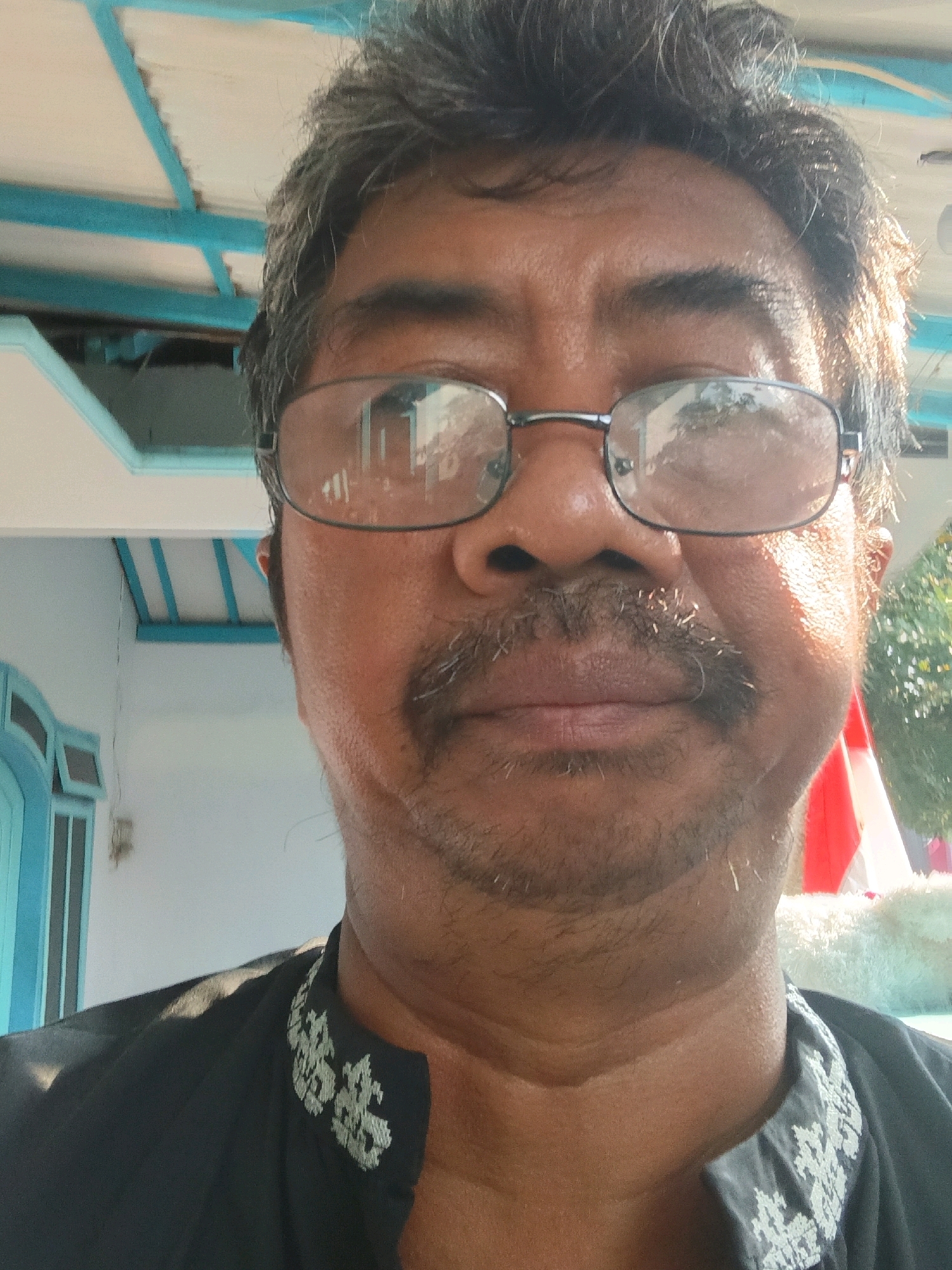 A close-up, front-facing, selfie-style shot shows a man with dark hair and a mustache, wearing glasses and a black shirt with white embroidered trim around the collar. He is standing outdoors, with a white building and blue trim visible in the background, suggesting the location may be a residence in Tanggul Wetan, Indonesia. The sun is shining, casting shadows and highlighting the man's face. A hint of red and white, possibly a flag or banner, is visible to the right of the man.