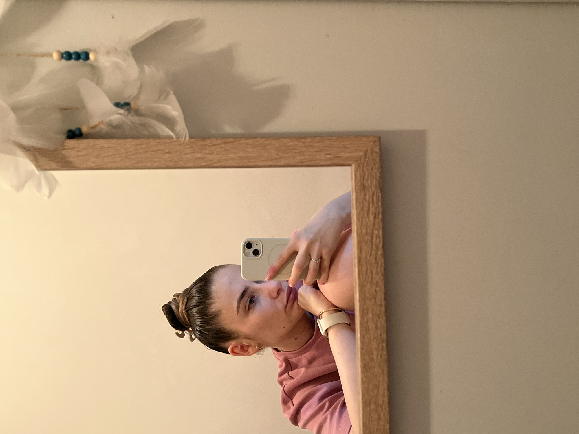 A young woman is taking a mirror selfie in an indoor setting. The image, if rotated 90 degrees counter-clockwise for standard viewing, shows her reflection in a rectangular mirror with a light wooden frame. She is captured from the chest up, looking towards her phone's camera with a neutral, slightly contemplative expression. Her dark hair is neatly pulled back into a bun. She wears a pink long-sleeved top.
She holds a light-colored smartphone, featuring two prominent camera lenses, in her left hand. Her right hand, adorned with a ring on her ring finger and a light-colored watch or bracelet on her wrist, rests near her chin. The background behind the woman in the reflection consists of light-colored walls; one is a soft off-white or light beige, and another to the right is a muted gray-beige. Above the mirror, to the upper left of the reflected scene, hangs a decorative item made of white, feathery material, possibly a dream catcher, strung with blue and light wooden beads. The lighting appears soft and even, suggesting either daytime or well-lit indoor conditions. There is no visible text in the image.