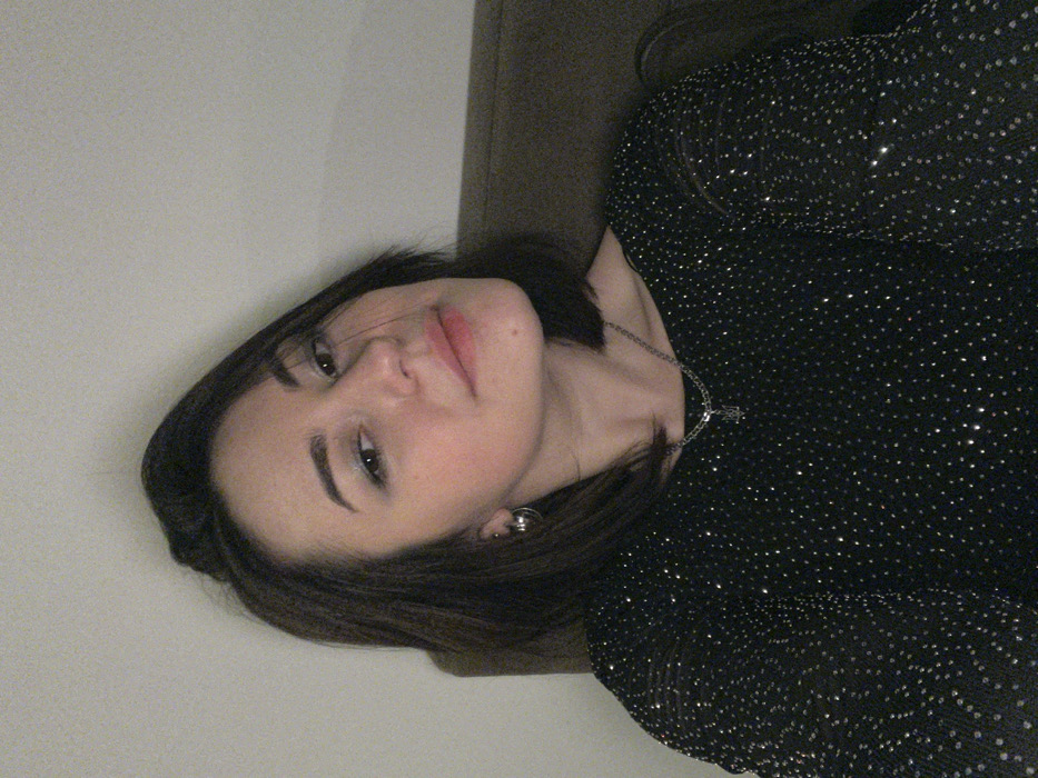 A portrait-style image depicts a single female individual positioned indoors. She has dark brown, medium-length hair, a light complexion, and is looking directly into the camera with a subtle smile. She wears a black long-sleeved top adorned with numerous small, reflective silver dots. Accessories include a silver-colored hoop earring on her left ear and a silver-colored chain necklace with a small trident-shaped pendant. The background consists of a plain, light-colored wall and the dark brown backrest of what appears to be a chair or sofa. The scene takes place in Budva, Montenegro.
