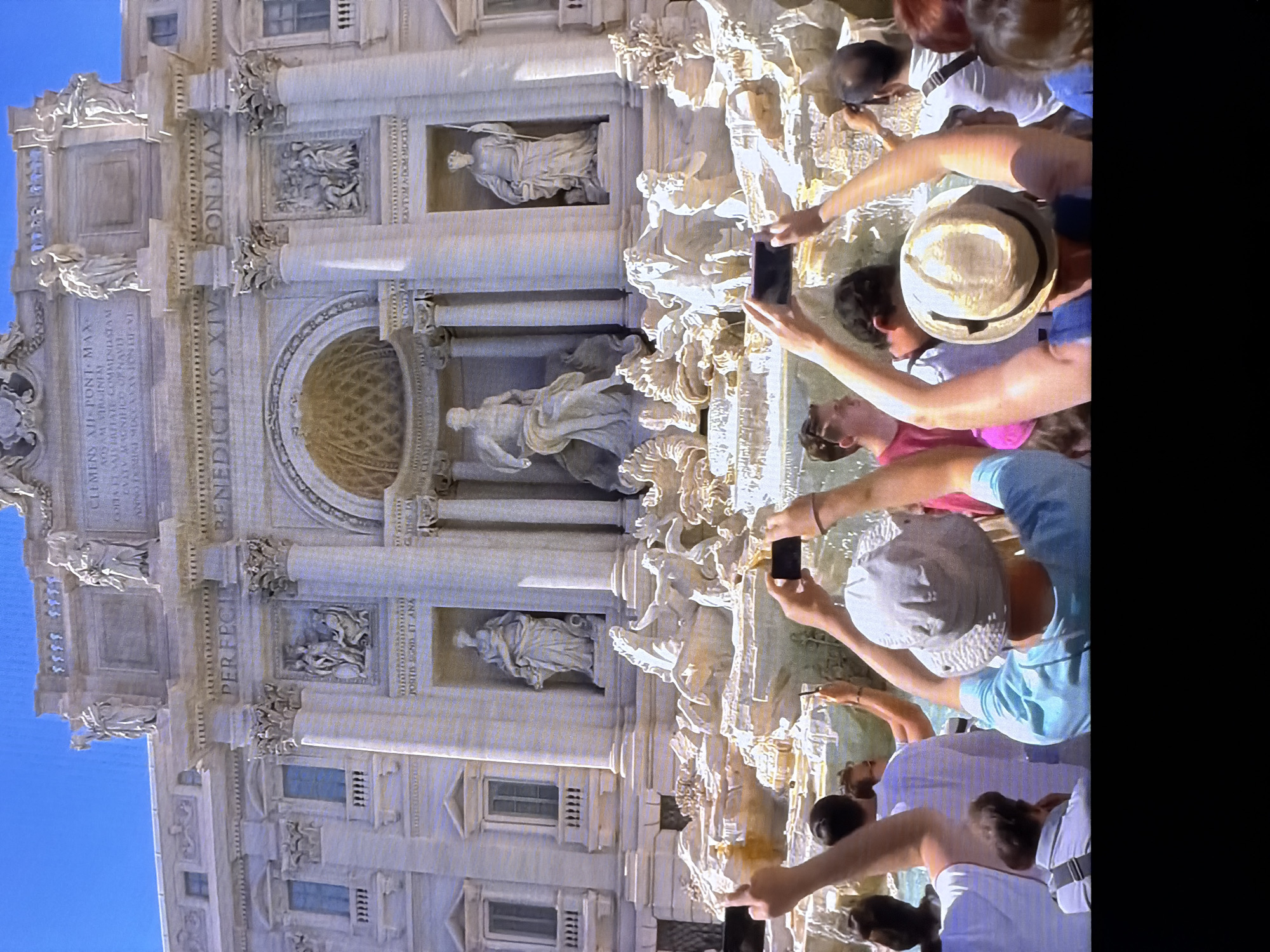 The image shows a group of people in front of a large, ornate fountain. Many of the individuals are holding up smartphones, appearing to take pictures or videos of the fountain. The fountain itself is a multi-tiered structure with sculptures and water features. The people are dressed in casual clothing, suggesting a tourist or public setting. The environment appears to be outdoors during the daytime, with a clear blue sky visible. There is no indication in the image that this scene is located in Chirchik, Uzbekistan.