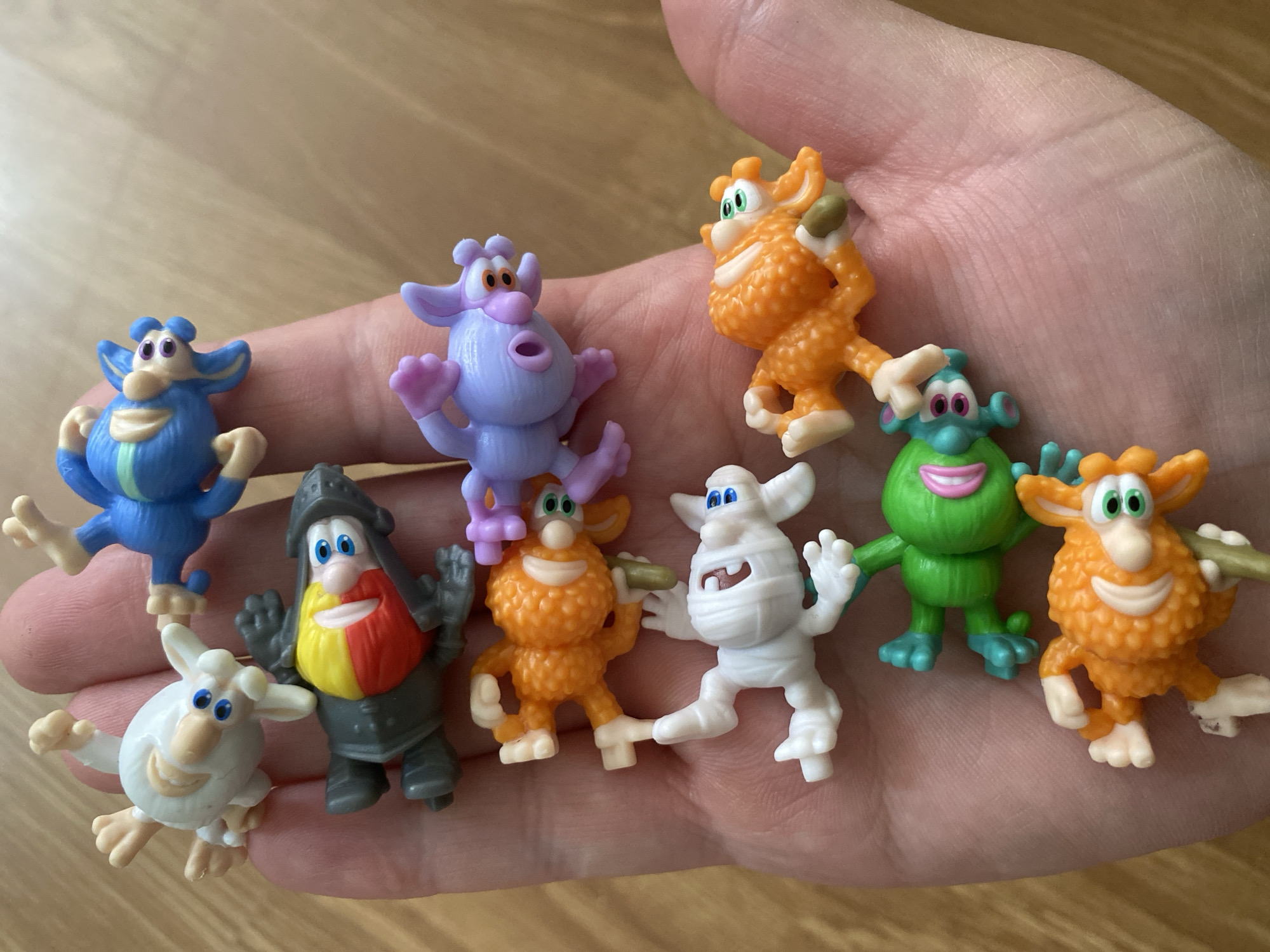 A hand is holding a collection of ten small, colorful plastic figures. The figures are diverse in color and design, including orange, purple, blue, green, white, and gray. The figures seem to be characters from a cartoon or children's show. The hand is resting on a wooden surface. The background is blurred. There is no indication that this photo was taken in Sochi, Russia.