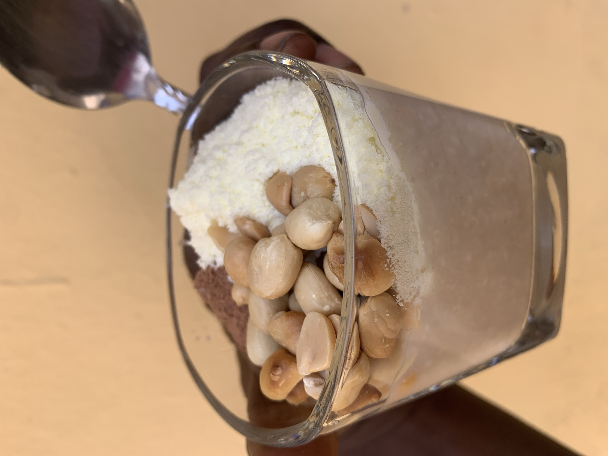A square glass dessert dish held by a person with brown skin is filled with layers. The bottom layer appears to be chocolate mousse. On top of that is a layer of what appears to be shredded white cheese, topped with light brown nuts. A light brown liquid fills the other half of the glass. A spoon is present above the glass. The setting appears to be indoors on a yellow-brown surface. There is no explicit evidence to indicate its location in Rafin Par, Nigeria, or any city, and therefore this cannot be confirmed.