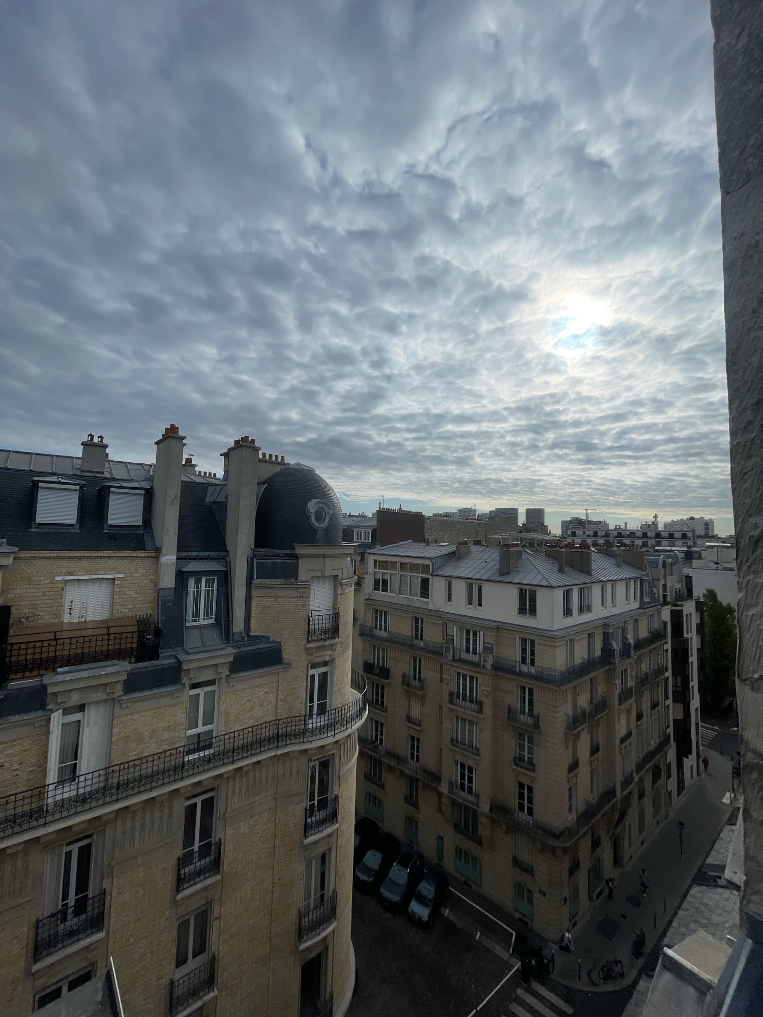 This image captures an elevated view of Parisian rooftops and street life under a cloudy sky. The architecture is typical of Paris, with mansard roofs, cream-colored brick buildings, and ornate balconies. A curved building in the foreground is particularly prominent, featuring multiple stories with arched windows and iron railings.
On the street below, several cars are parked. Pedestrians are visible, some walking along the sidewalk and others crossing a pedestrian crossing. The overall atmosphere suggests a slightly overcast day in a residential area of Paris. The sun is partially visible through the clouds, creating a diffused light. The image appears to have been taken from a high vantage point, possibly a window or rooftop.