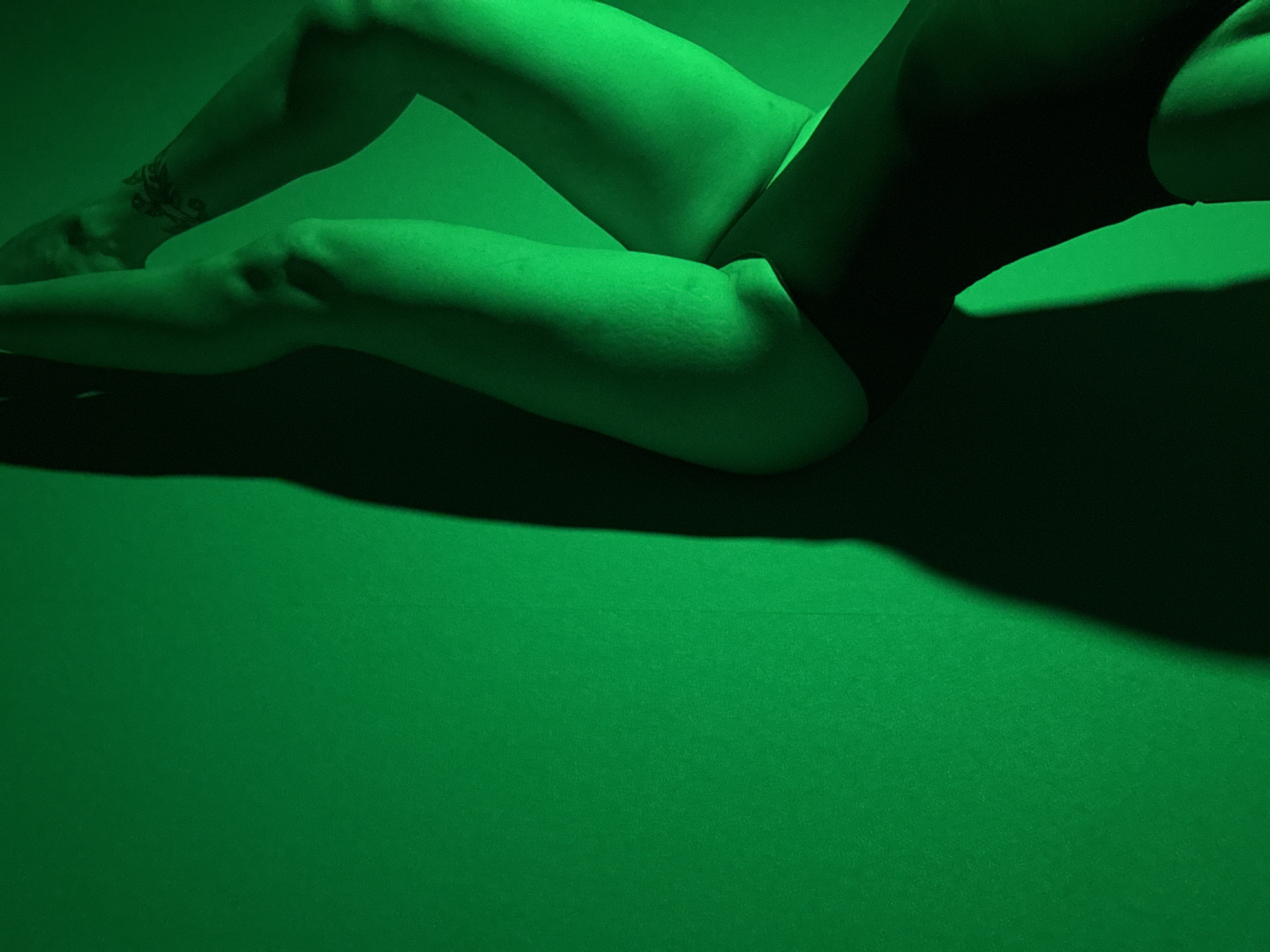 A photograph, predominantly green in tone, depicts a person lying on a green surface. The individual's body is partially illuminated, showcasing a leg with a visible tattoo. The scene's stark green coloration and the person's pose dominate the composition. There is no evidence to suggest a location.
