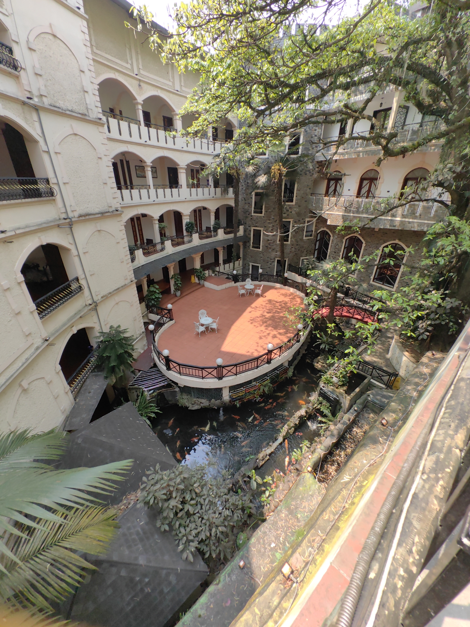 The image depicts an enclosed multi-story courtyard within a building in Sa Pa, Vietnam, viewed from a high angle. The building's architecture varies: the left section features a light-colored, cream-toned facade with arched balconies and windows, and black-and-gold wrought iron railings. The right section displays a darker, grey stone-textured facade with arched openings and dark railings.
A raised, oval-shaped patio with terracotta-colored tiles occupies the courtyard's center, bordered by a black metal railing with white spherical lights. Two white round tables, each with four chairs, are arranged on the patio. Below the patio, a clear koi pond contains numerous orange, white, and dark-colored koi fish. Abundant green vegetation, including large moss-covered trees, palm-like plants, and shrubs, are arranged around the pond and building levels. A small, arched red bridge spans a section of the pond on the right. Dark, pyramidal roof structures are visible in the lower-left, adjacent to the water. Natural sunlight illuminates the scene, casting shadows. No individuals are visible.