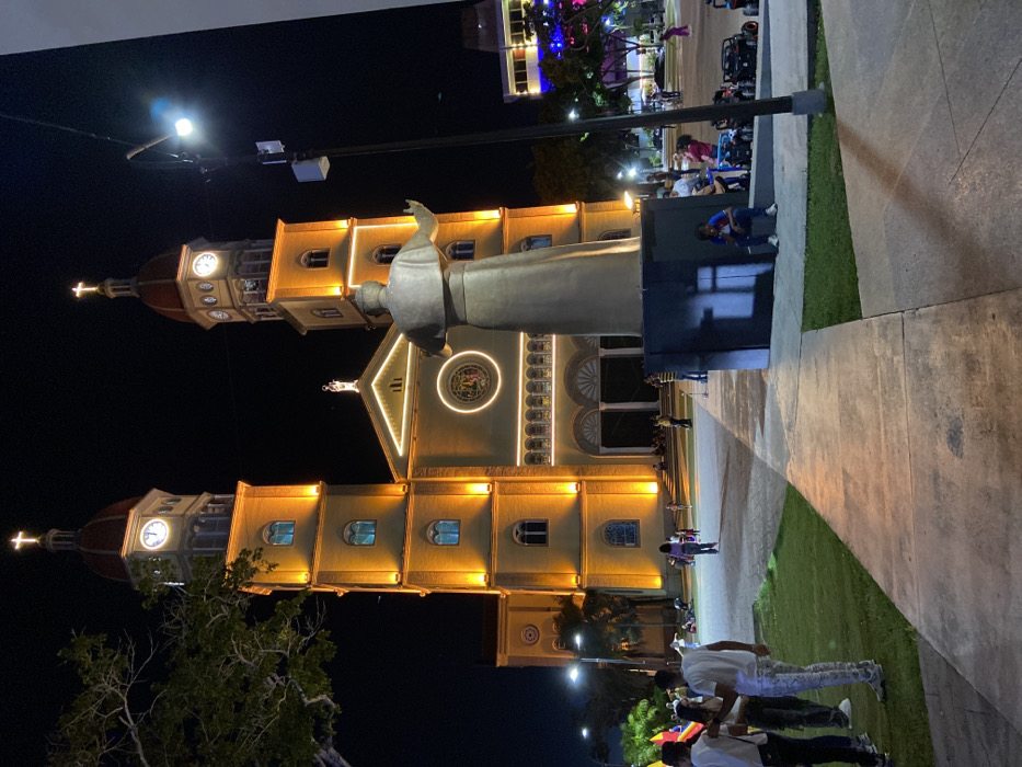 A church building with twin towers is illuminated at night. A statue of a figure in robes stands in front of the building. The structure is decorated with lights, particularly a circular pattern above the entrance. Several people are gathered in the foreground on a walkway. Trees and additional lights are visible in the background, indicating a populated or public area.