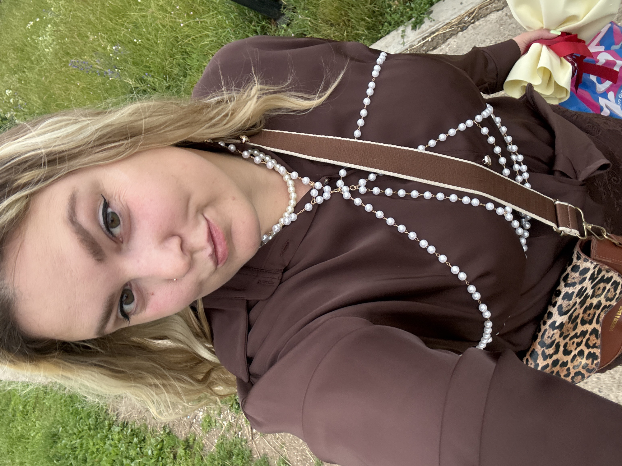 In Марковский, Russia, a blonde woman with light skin and dark eyebrows is pictured from the chest up. She is wearing a brown button-down shirt with a decorative pearl body chain. A brown crossbody bag with a leopard print flap is visible at her hip. The background consists of green grass and a path. The woman is holding a wrapped gift with a yellow ribbon.