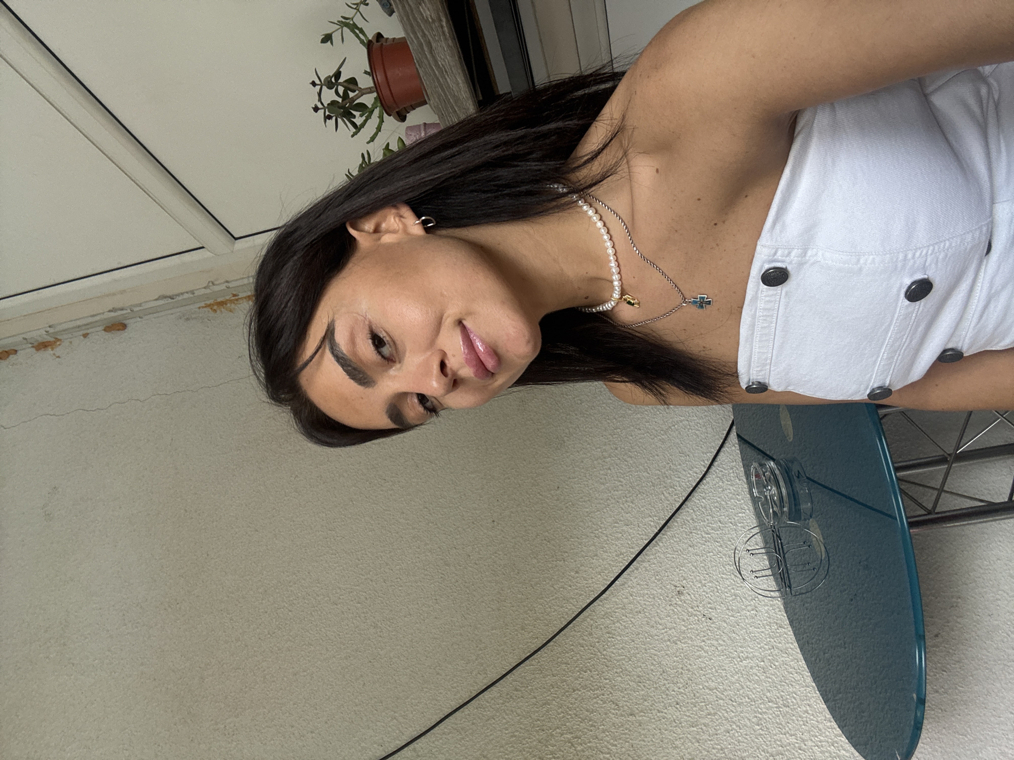 A young woman with dark hair and brown eyes is pictured from the chest up, outdoors on a balcony. She is wearing a white strapless top and two necklaces, one with pearls and the other with a cross pendant. She is looking at the camera and smiling slightly. In the background, a small round table with an ashtray is visible, as well as a potted plant on a shelf. The wall behind her appears to be textured and has some peeling paint in places. The overall lighting suggests it is daytime.