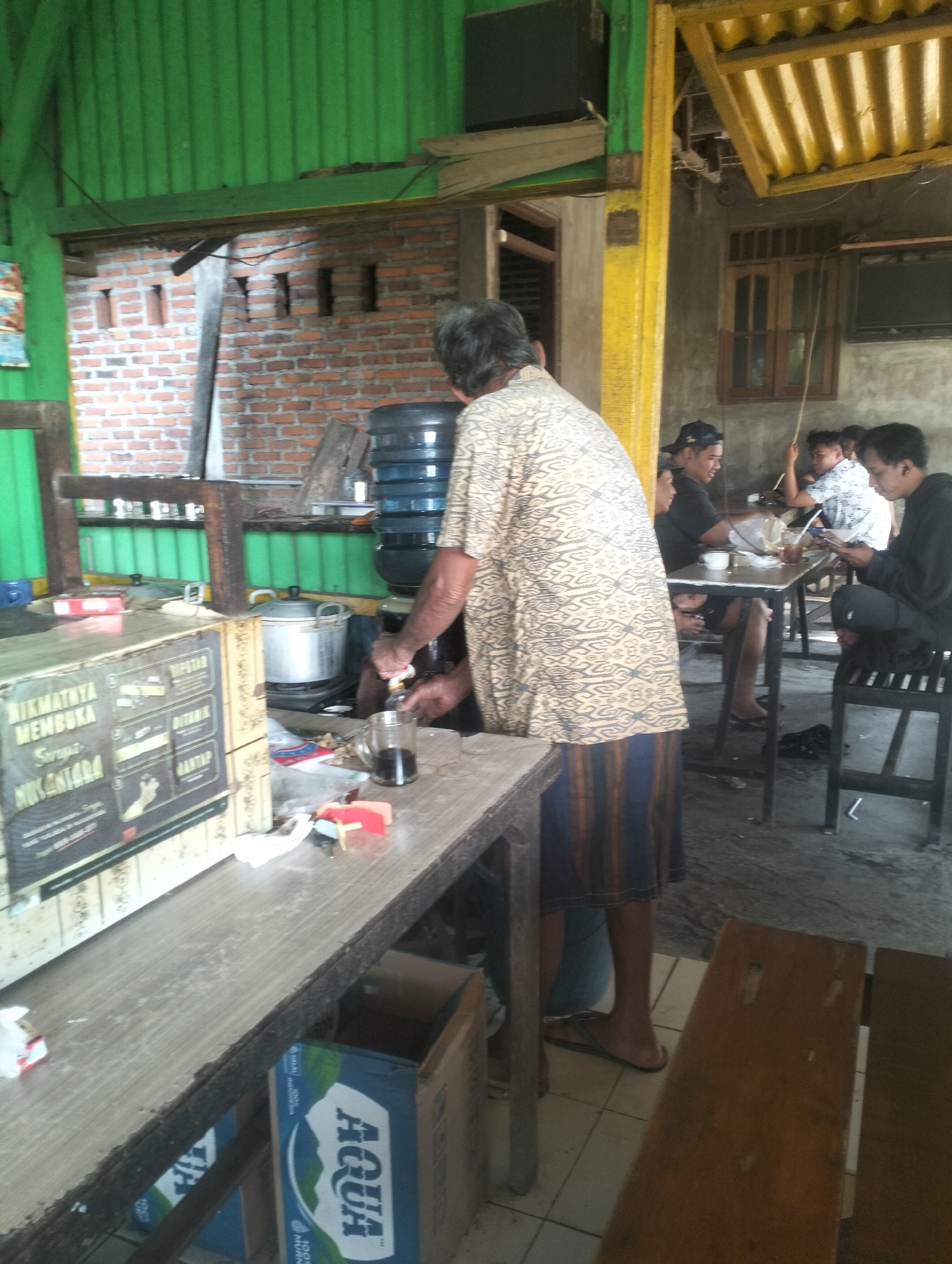 The image depicts an outdoor food stall or small eatery. In the foreground, a man wearing a patterned shirt and striped skirt is pouring a dark liquid from a bottle into a glass mug. He stands behind a long wooden counter. On the counter are several items, including a pot with a lid, a water dispenser, and a box with text and images. To the left, a sign with Indonesian text is affixed to the counter. In the background, several other people are seated at tables, some of whom appear to be eating or drinking. The environment is rustic, with brick walls and a corrugated metal roof visible.