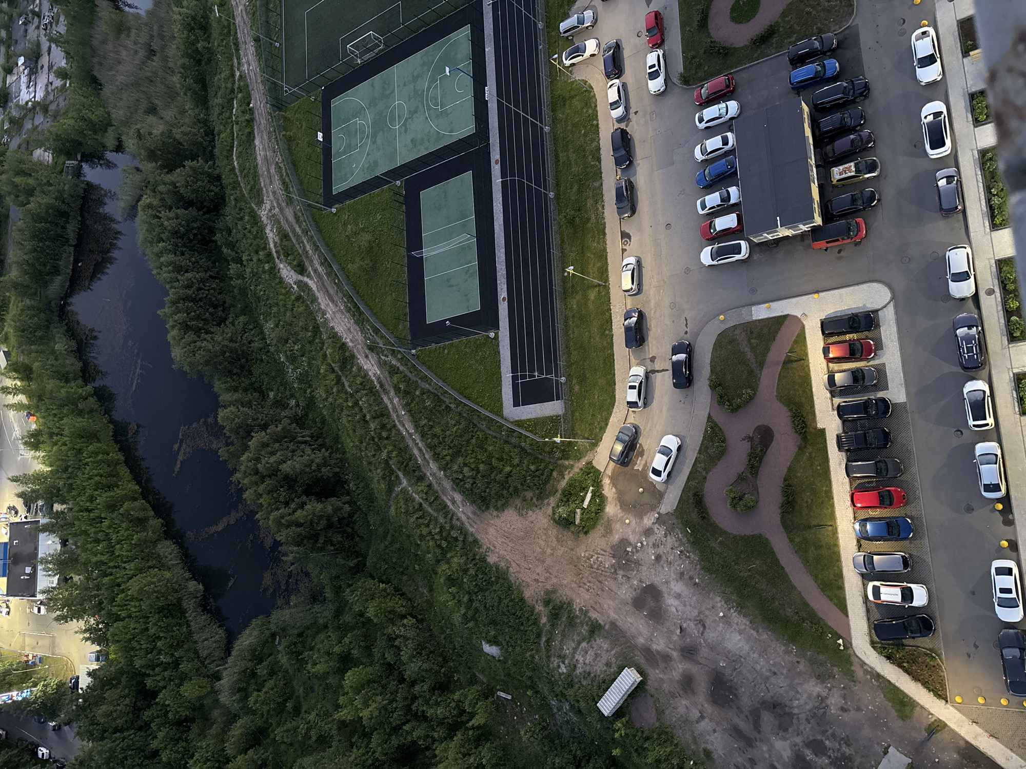 The aerial photograph, taken in Unknown, Unknown, depicts an urban outdoor environment characterized by a body of water, dense vegetation, sports facilities, and a residential area with parked vehicles. In the upper left quadrant, a dark, irregular-shaped body of water is surrounded by a thick line of trees and reeds. Adjacent to this natural feature, a sports complex is visible, featuring a basketball court with white markings and a smaller rectangular court with a net, possibly for tennis or volleyball. Beyond these courts is a fenced-off area with what appears to be a soccer field, the grass a vibrant green.
A network of unpaved roads and paths winds through the landscape, some showing signs of tire tracks and erosion. In the foreground and extending towards the bottom of the image, a residential area is evident. Numerous cars are parked in designated lots and along the roadside. The vehicles vary in color and model, including sedans, SUVs, and what appears to be a white delivery truck or trailer. The architecture of the residential buildings is not clearly discernible, but a low-rise structure with a dark roof and yellow walls is positioned near the sports facilities.
The lighting suggests it is either early morning or late evening, with soft shadows cast by the objects in the scene. The overall impression is a blend of urban development and natural elements, with recreational spaces integrated into the residential landscape. No people are visible in the photograph.