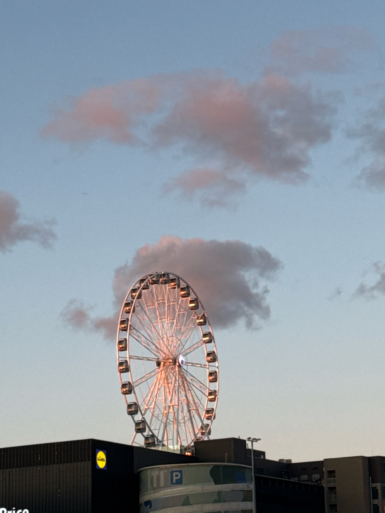 The image captures a Ferris wheel, illuminated by warm lights, against a pale blue sky with scattered pinkish clouds. The Ferris wheel is positioned to the right of the frame, its metallic structure and gondolas clearly visible. Below the Ferris wheel, a dark building with a prominent yellow circular logo, which reads "Lidl", is visible on the left side of the frame. To the right of the Lidl building, another building with a blue and white sign featuring the letters "T I P" is partially visible. The lighting suggests it is either sunrise or sunset. The scene appears to be an urban environment, with the Ferris wheel likely being an amusement attraction or part of a city skyline. The image is likely taken in Tallinn, Estonia, where a Ferris wheel known as the "Tallinn Eye" is located near the harbor and has Lidl supermarkets in its vicinity. There are no discernible people or specific actions taking place.