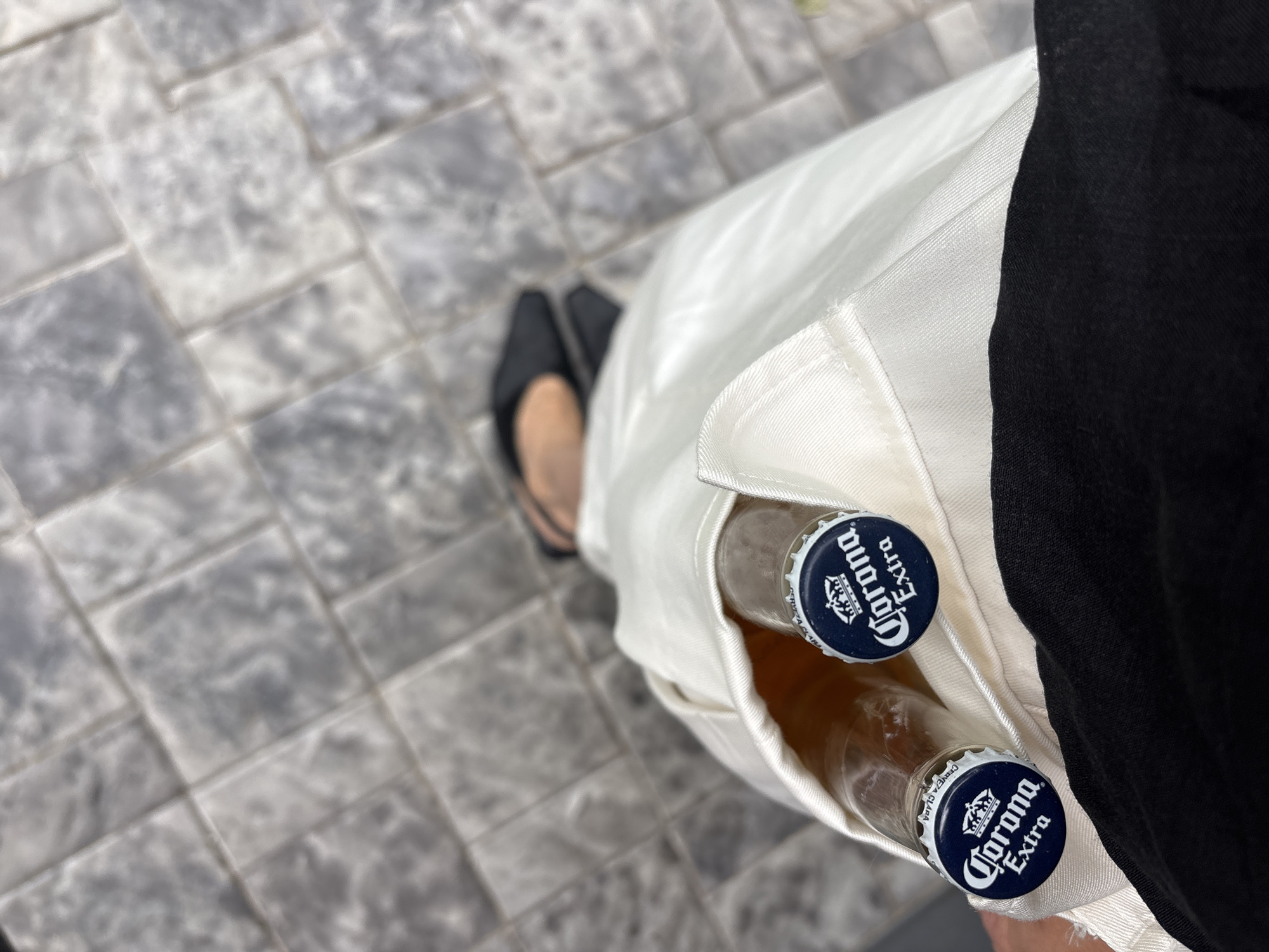 In a close-up, overhead shot, two Corona Extra beer bottles are held within the white fabric of what appears to be a garment, possibly the pocket of trousers or a coat. The dark blue caps of the bottles, featuring the distinctive Corona Extra logo with a crown, are visible. The bottles are transparent, showing a small amount of liquid at the bottom.
In the background, a person's feet are visible, wearing black slip-on shoes. The ground beneath is paved with light gray square tiles, arranged in a diagonal pattern. The tiles have a slightly textured and worn appearance, suggesting an outdoor urban setting. The overall lighting is bright, indicating daytime. The specific location in the city of Chaikovsky, Russia, cannot be determined from the visual information provided in the image.