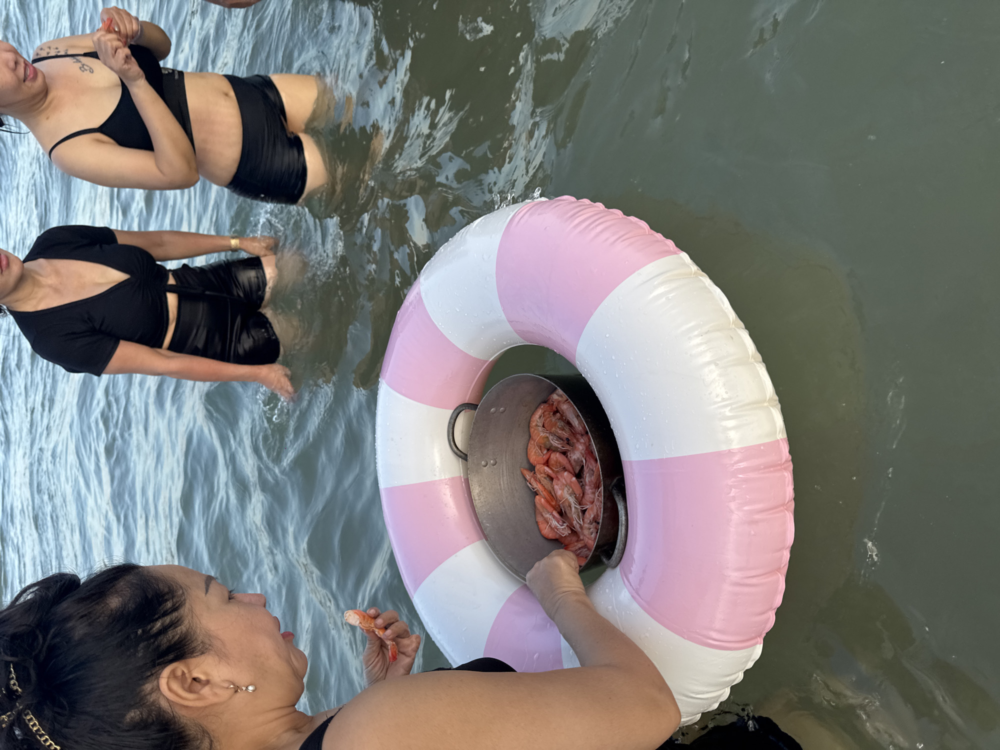 The image depicts three women standing in shallow, murky water, likely a river or bay, in Thuận Thành, Vietnam. One woman in the foreground, with her hair pulled back in a bun and wearing a black swimsuit, holds a cooked shrimp in her right hand and is reaching into a metal pot filled with more cooked shrimp. The pot is resting inside a pink and white striped inflatable ring. To her left, another woman, also in black swimwear, stands facing away from the camera. To the right of the foreground woman, a third woman, wearing a black bikini top and black shorts, looks down at her hands and is holding a shrimp. The water is calm with ripples, and the lighting suggests it is daytime. The scene appears to be a casual gathering or outing.