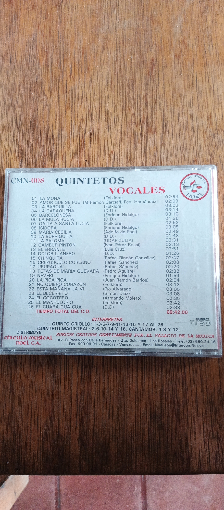 This is a close-up, eye-level shot of a CD case, likely displayed on a wooden surface. The CD case is primarily white with text and graphics printed on it. The text is in Spanish and lists song titles, artist names, and timings. Some of the songs are labeled as "Folklore" or have composer attributions. There's also information about the distributor, "CÍRCULO MUSICAL DOEL C.A.," and their contact details in Caracas, Venezuela. A circular logo with the words "Circulo Musical DOEL" is visible on the right side of the case, along with the "COMPACT disc" logo. The overall scene appears to be a retail display or personal collection of music. The wooden surface beneath the CD case has a warm, natural tone with visible wood grain, suggesting it might be a shelf or table. The lighting is even, with no strong shadows, indicating a well-lit indoor environment, possibly a music store or a home. The image focuses on the CD case as the main subject, highlighting the details of the music collection.