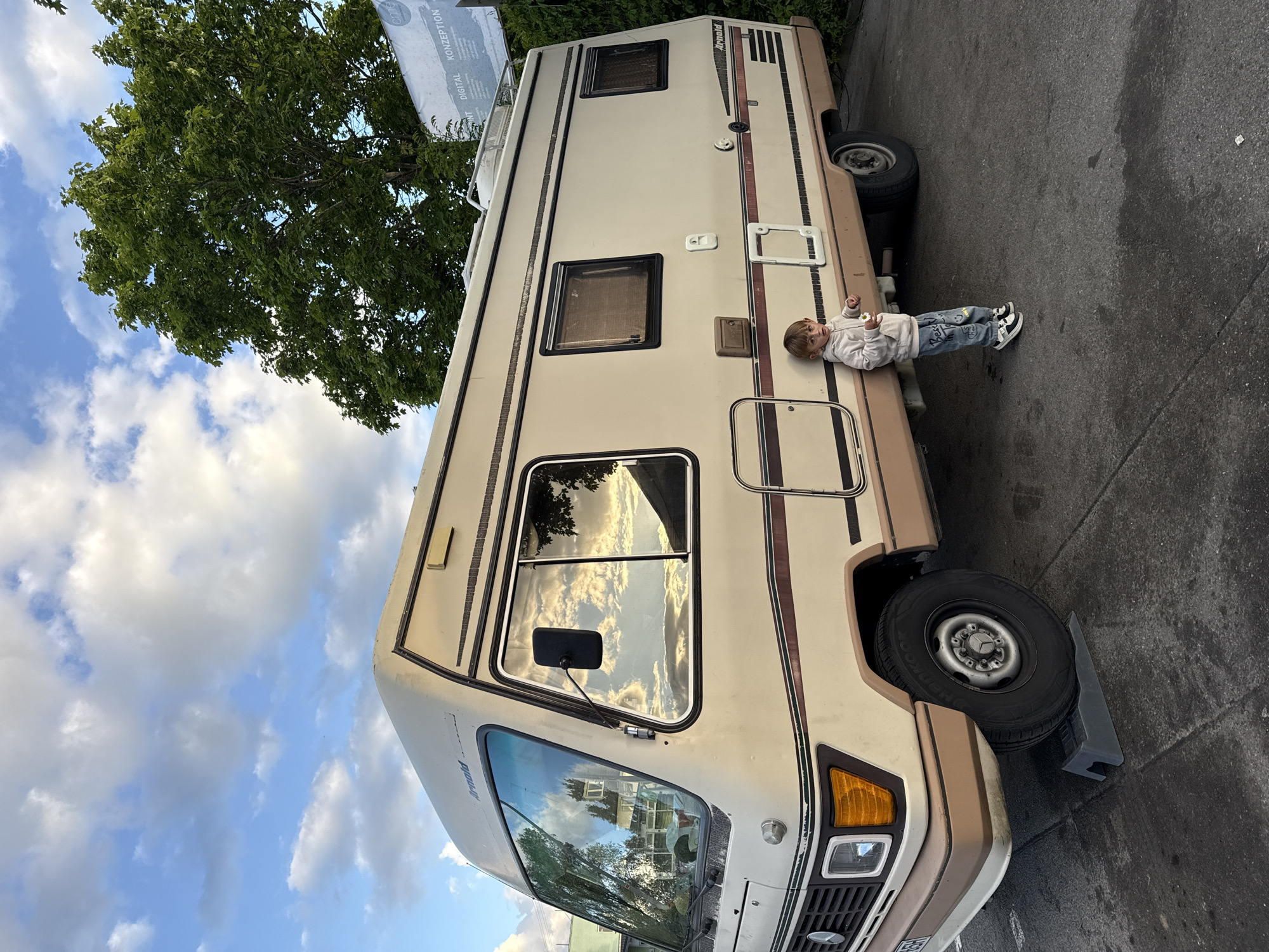 A photograph captures a toddler resting on the side of a camper van. The child, dressed in a light-colored sweatshirt and jeans with black and white shoes, is lying horizontally against the van. The camper is beige with brown trim and several windows. Reflections of the sky and trees are visible in the van's windows. A large tree with dense green foliage overhangs the top left of the frame, while a partially obscured sign is located behind the tree. The ground is paved, and there's a wheel chock next to the front tire of the camper. The photograph appears to have been taken in Riedlingen, Germany.