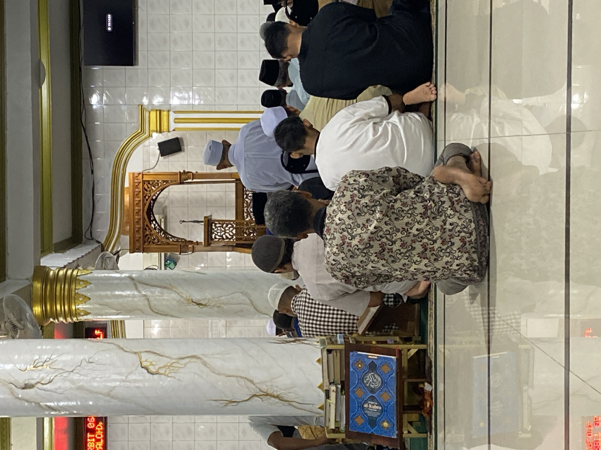 The image depicts an interior view of a mosque in Lhokseumawe, Indonesia, during prayer. Several men are seated on the floor, facing the front of the mosque, suggesting they are engaged in prayer. The individuals are dressed in traditional attire, with many wearing skullcaps. In the foreground, a man in a patterned shirt is seen from behind, with his feet bare. To his left, a stack of books rests on a wooden stand, with an open book displaying Arabic script in blue.
The mosque's architecture is visible, featuring large, decorative pillars with a marble-like finish and gold accents. A wooden minbar, the pulpit, is situated at the front of the prayer hall. The walls are tiled in white, and a television screen is mounted high on the wall, displaying the time "12:14:24". The floor is a polished, reflective tile, creating reflections of the scene. The overall lighting suggests it is either early morning or late afternoon. The presence of prayer mats and the arrangement of the attendees indicate a religious gathering.