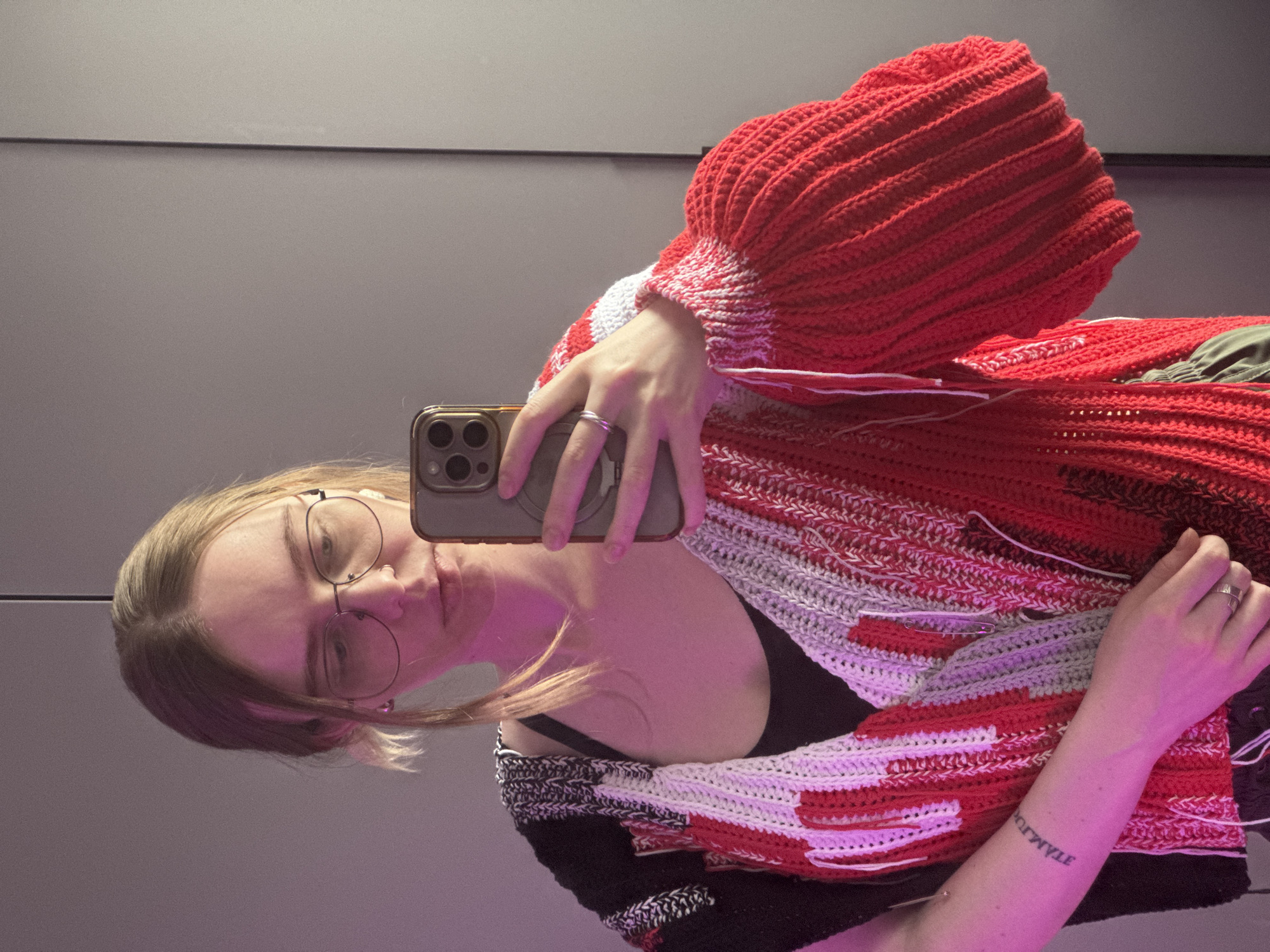 A woman with light skin, light brown hair, and glasses is taking a selfie in a mirror. She is wearing a large, red, white, and black crocheted sweater or cardigan over a black top. The sweater has exaggerated sleeves. The woman is holding a gold iPhone in her right hand, capturing her reflection. A silver ring is visible on her right ring finger. Her left arm is also visible, showing a tattoo of what appears to be Cyrillic text. The background behind her is a smooth, gray wall. No information in the image suggests the location is in Krasnoyarsk, Russia.