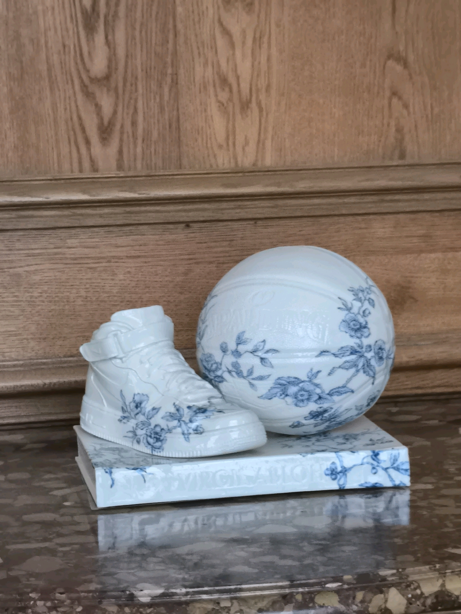 A white, porcelain decorative sneaker with blue floral patterns sits atop a white, porcelain book with blue floral patterns. Beside the book and sneaker is a white, porcelain basketball with similar blue floral patterns. The objects are displayed on a polished marble surface in front of a wooden paneled wall. The scene is indoors, with no people visible. The lighting suggests an interior setting. The objects are likely decorative art pieces.
