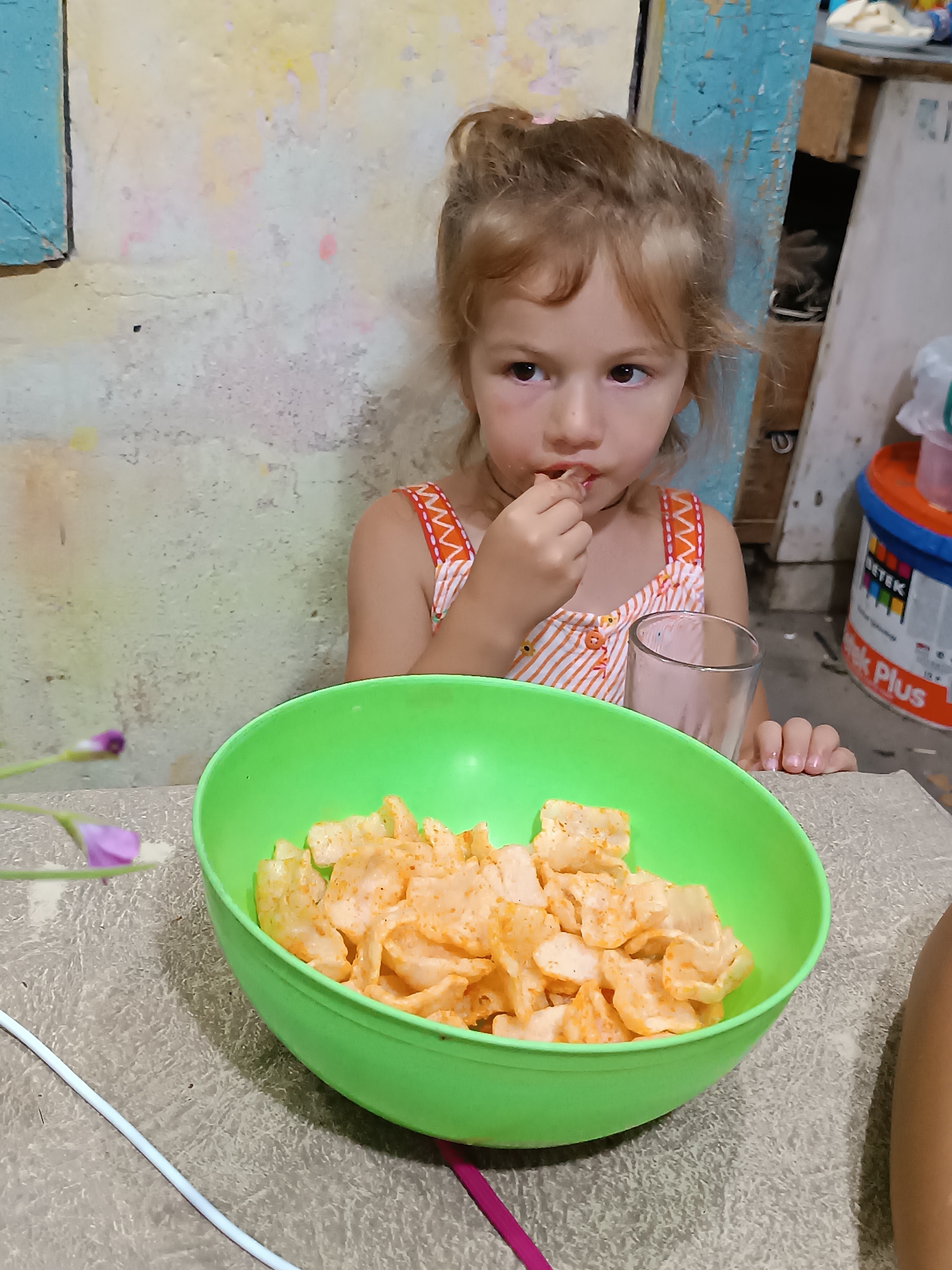 The image depicts a young girl, likely Caucasian, with blonde hair pulled into a bun, sitting at a table. She is wearing a white and orange striped sleeveless dress and is in the process of eating. In front of her is a large, bright green plastic bowl overflowing with what appear to be puffed snacks, possibly chips or crackers, with an orange seasoning. To her right, a clear glass with a light-colored liquid, potentially milk or a shake, is placed on the table. Her hands are visible, with her right hand near her mouth, and her left hand resting on the table. The background suggests an indoor setting with a rough, textured wall painted in muted tones of yellow and beige, with patches of lighter paint and some darker marks. To the right, a blue, distressed wooden structure is visible, and behind that, a large industrial-style bucket, possibly for paint, is present. A small purple flower with green leaves is positioned on the table to the left of the bowl. The overall environment appears to be modest and possibly rustic. The image was taken in Khashuri, Georgia.