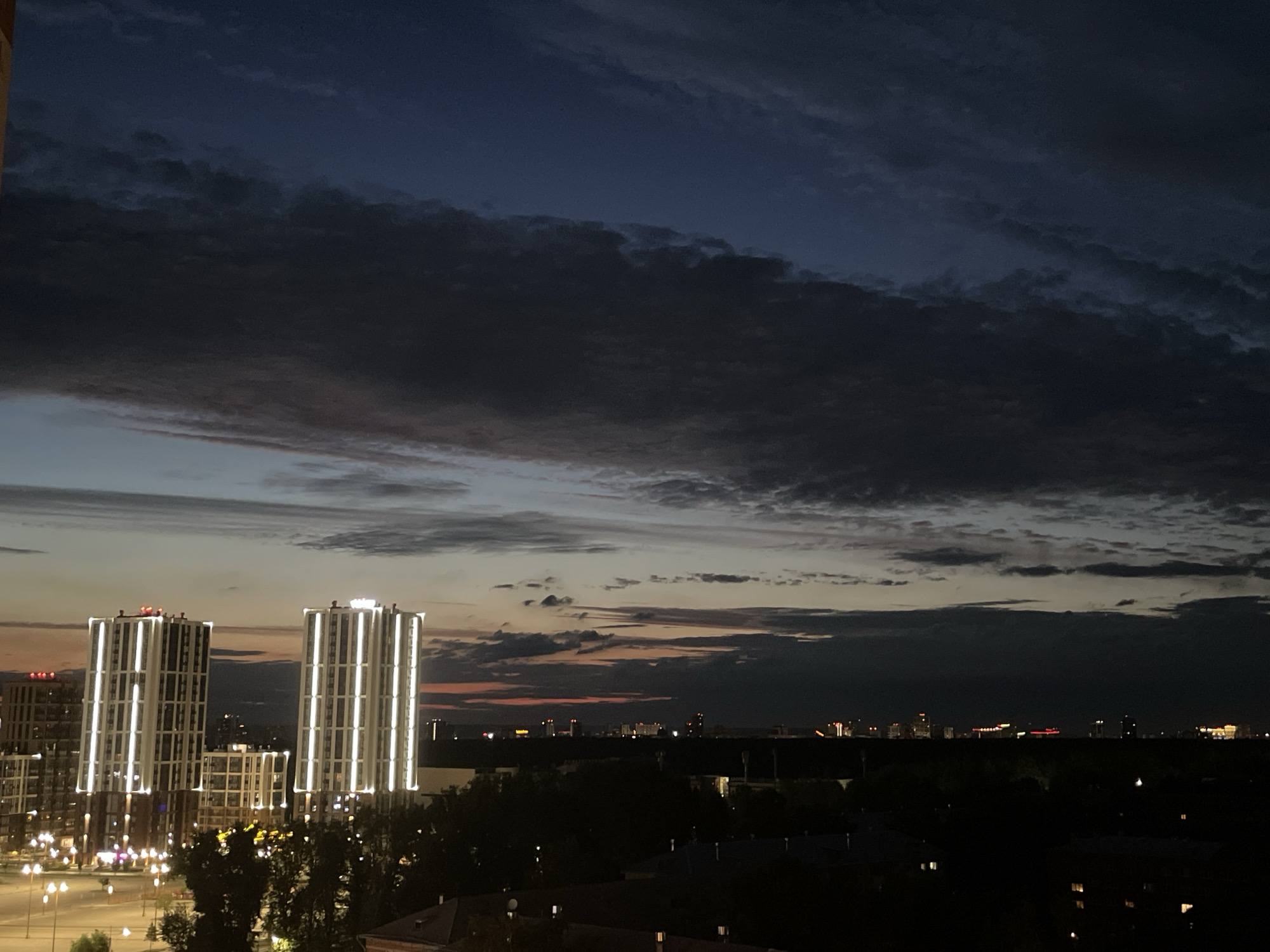 The image captures a twilight cityscape in Zarechnaya (Gamovskoe S/P), Russia. Dominating the foreground are two tall, modern buildings illuminated with vertical strips of white light. To the left of these, a portion of another multi-story building is visible. In the middle ground, a line of trees obscures a distant city skyline dotted with lights. The sky above is a dramatic blend of dark blues and grays, with scattered clouds and a faint band of orange and pink near the horizon, indicative of sunset or sunrise. The overall impression is of an urban environment at dusk or dawn.