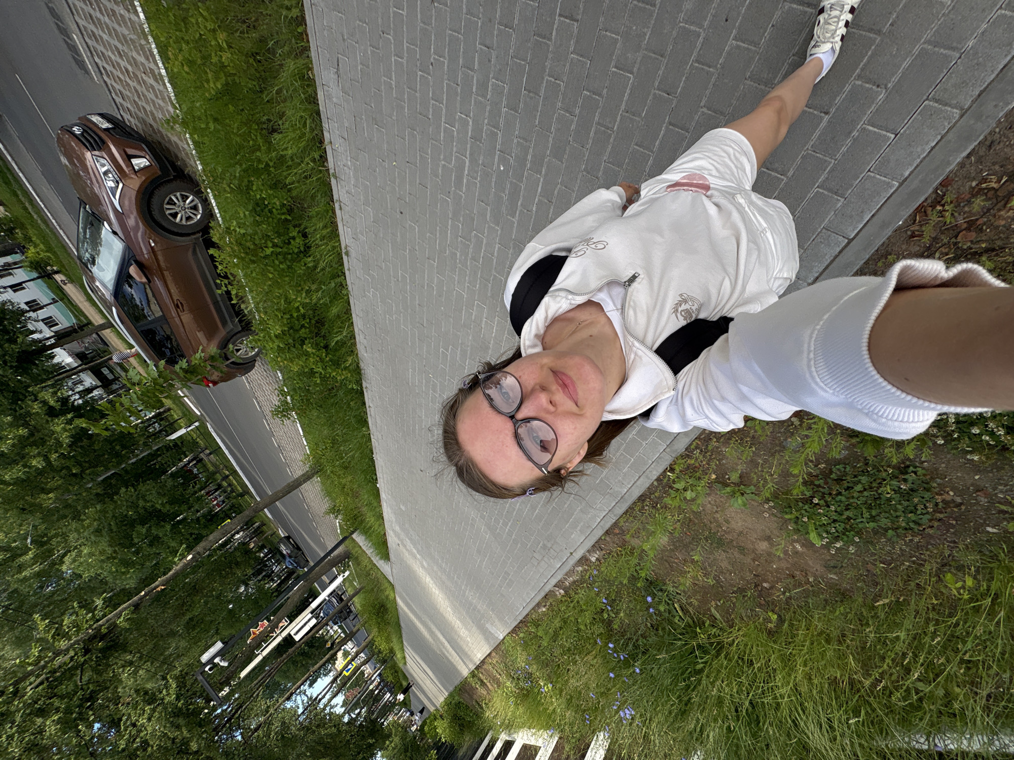 A young woman wearing glasses, a white hooded jacket, and shorts is taking a selfie on a paved sidewalk. She has a backpack on. To her left is a strip of green grass and trees. To her right is a road with a brown Hyundai car parked on the side, and in the background, there are buildings and more trees. The scene appears to be in Chaikovsky, Russia, given the presence of Cyrillic text on a street sign visible in the distance.