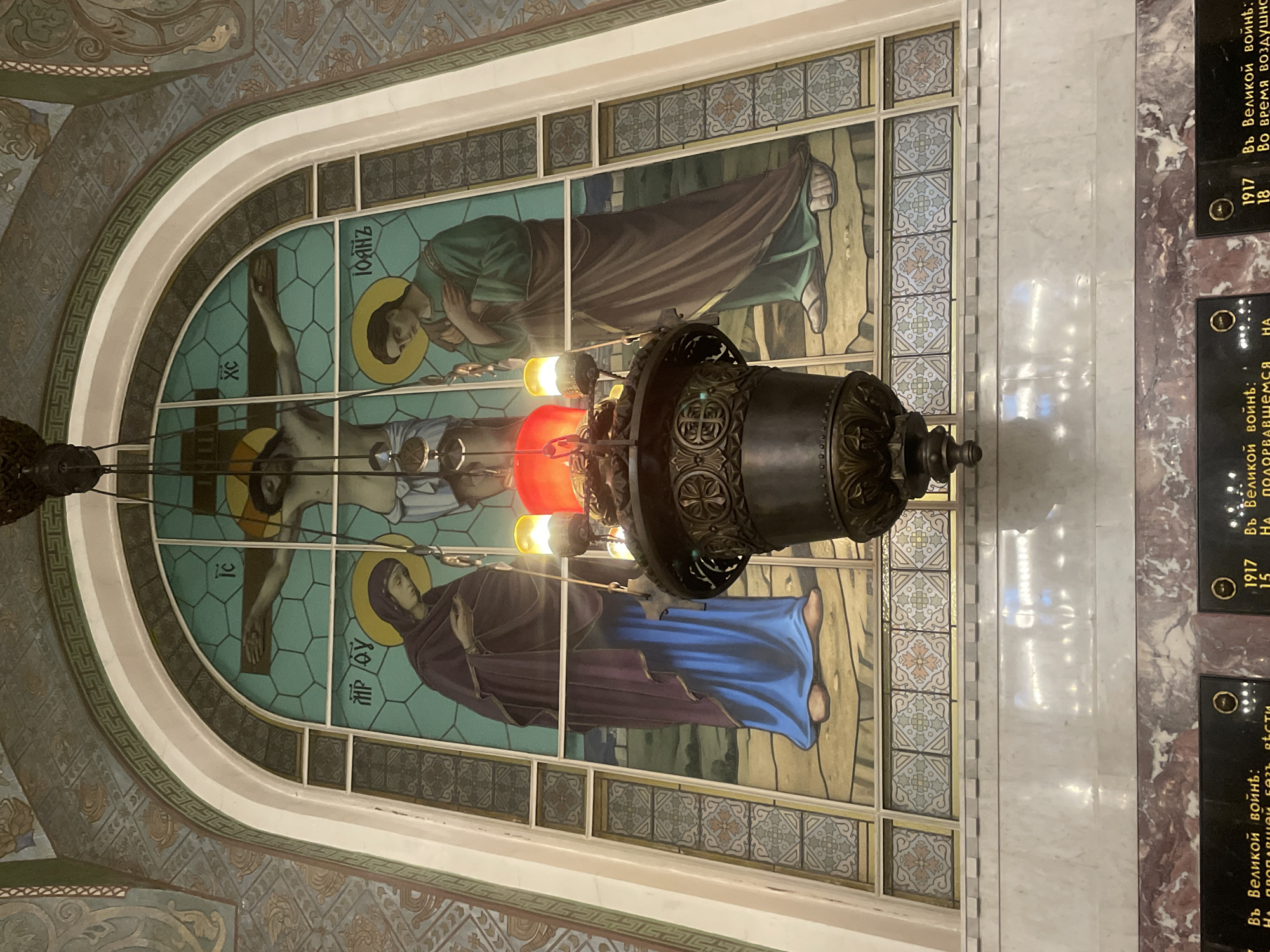 The image depicts the interior of a religious building, likely a church, featuring a large stained-glass window depicting the crucifixion of Jesus Christ. Christ is shown centrally on the cross, with the Virgin Mary on his left and John the Evangelist on his right. Above their heads are Greek letters likely representing their names or titles. The figures are rendered in a traditional religious style.
A decorative, ornate metal chandelier hangs in the foreground, obscuring a portion of the stained-glass window. The chandelier has multiple lights, some of which are illuminated with red and yellow hues. The stained-glass window is set within an arched frame decorated with patterned tiles and a Greek key motif. The ceiling above the window also features patterned artwork.
Below the stained-glass window is a marble structure, part of which appears to be a memorial. Black plaques with inscribed text are visible at the very bottom of the image, suggesting this is a commemorative space. The style of the architecture and the memorial plaques, particularly any visible dates or inscriptions, would be crucial in identifying the specific location.
Given the context of the Kronstadt Naval Cathedral, this scene likely represents a section of the cathedral's interior, possibly near the memorial to those who perished in World War I or other conflicts, as indicated by the partially visible text on the plaques. The cathedral, a prominent landmark in Kronstadt, Russia, is known for its rich interior decoration and historical significance.
