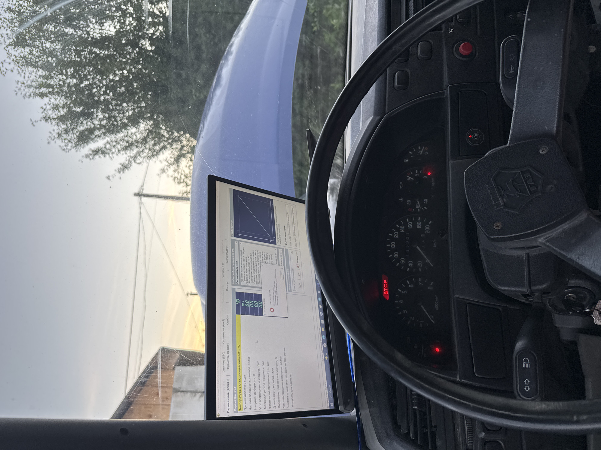 The image shows the interior of a vehicle, likely a car, with a laptop displaying diagnostic software open on the dashboard. The laptop screen shows various parameters related to engine diagnostics, including engine coolant temperature, crankshaft position, and throttle position. The dashboard features a steering wheel, instrument cluster with gauges for speed and RPM, and various indicator lights, one of which is red and labeled "STOP". The hood of the vehicle is open, revealing a blue engine bay. The scene appears to be outdoors, with a tree and a building visible through the windshield. The sky suggests it may be late afternoon or early evening. The image is taken from the driver's perspective. Based on metadata analysis, the image was geotagged in Krasnokamsk, Russia, suggesting the vehicle is located there. No people are visible in the image.