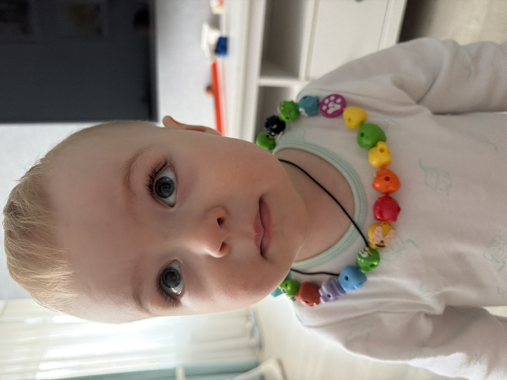 A close-up shot presents a baby, likely around 1-2 years old, with light blonde hair and striking blue eyes, adorned with a colorful beaded necklace. The necklace features various shaped and colored beads, including some with animal designs, strung on a black cord. The baby is wearing a light-colored, long-sleeved shirt with a light green-colored trim around the neck. The baby's gaze is directed towards the viewer, and the composition suggests the photograph was taken from a slightly elevated angle. The background is blurred, but elements of a room are discernible, with a white shelf visible on the right and a dark object, possibly a television, on the left. The scene appears to be indoors, possibly a residential setting. There is nothing to suggest that the location is specifically in Votkinsk, Russia, or any other particular city.