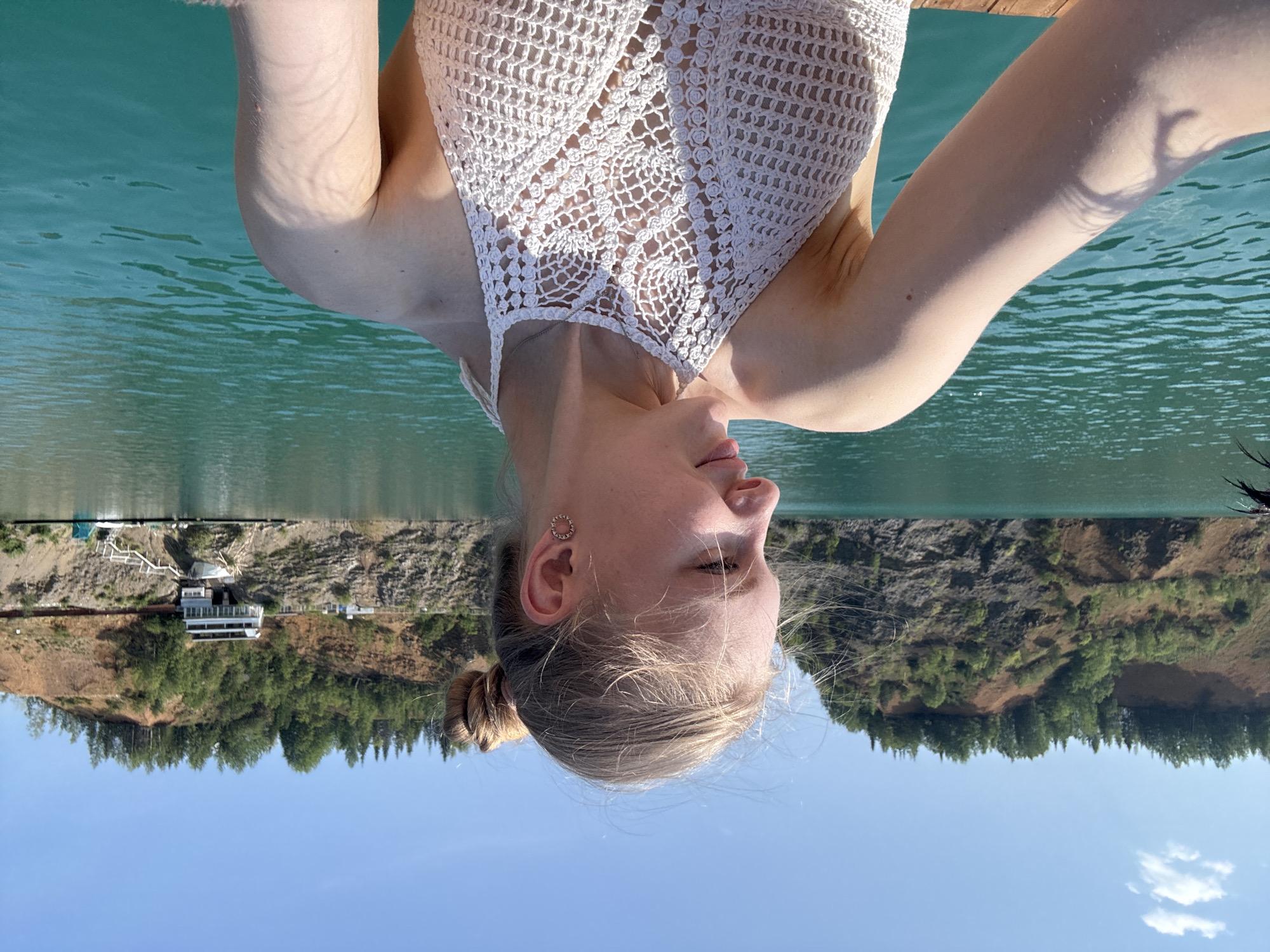 The image depicts a young woman from the chest up, posing on a wooden dock next to a body of clear turquoise water. She is wearing a white crocheted halter top. Her hair is pulled back into a bun, and she has a small, circular earring in her visible ear. Her head is tilted downwards, and her eyes are closed, suggesting a moment of relaxation or enjoyment.
The scene is set on a sunny day, with a clear blue sky visible in the background. The water appears calm and reflects the sunlight, creating a serene atmosphere. Behind the woman and the water, a steep, rocky hillside covered in lush green trees rises up. A white building with multiple windows and a few smaller structures are nestled on the hillside, possibly indicating a resort or residential area. The overall impression is one of a peaceful summer day in a natural, scenic location. The image appears to be taken from a low angle, possibly from within the water or from a boat.