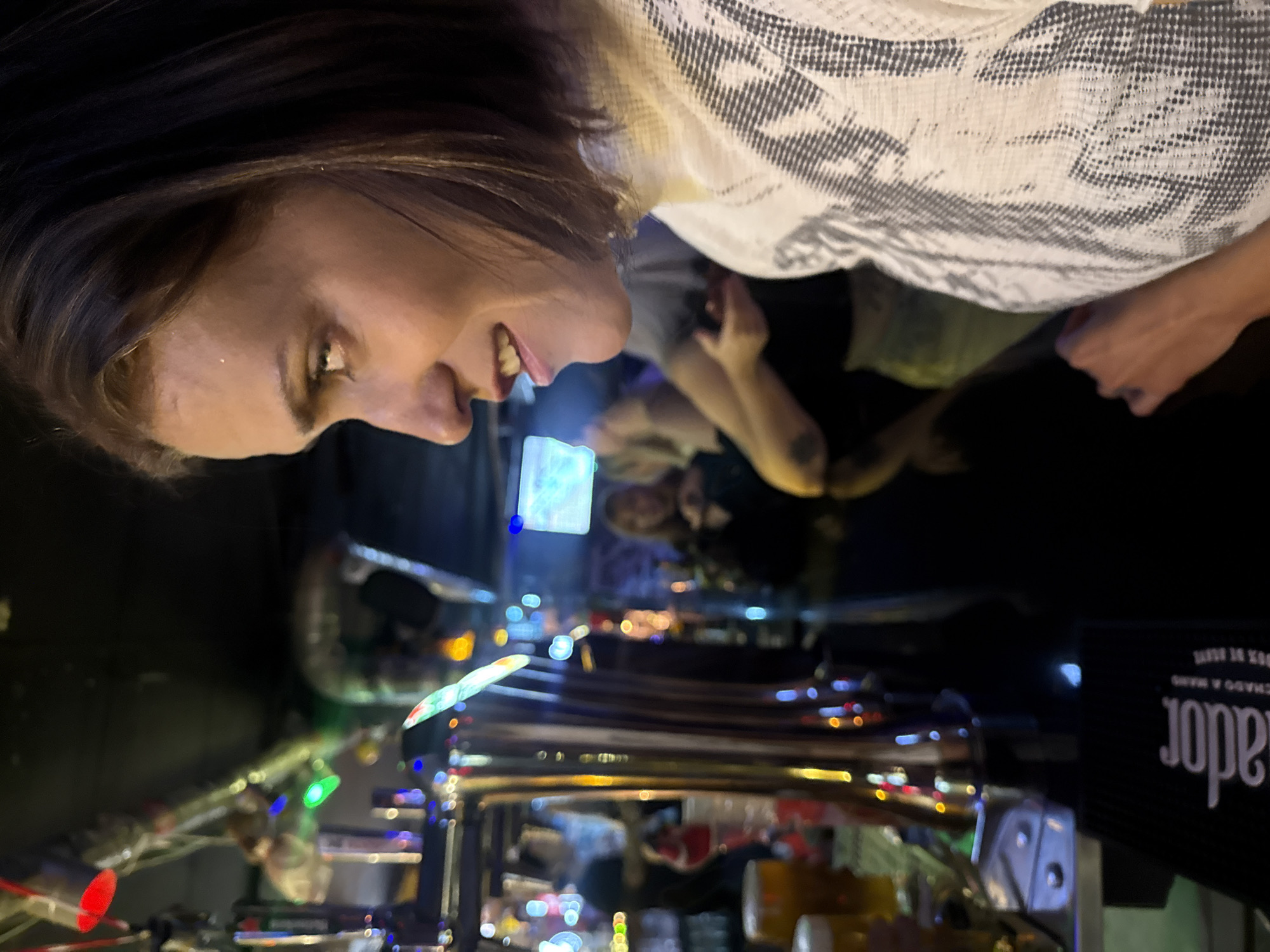 A woman with short brown hair and a patterned white shirt is looking at a beer tap in a bar. The beer tap is shiny metal with colorful lights. Behind the woman, other people are sitting at the bar. The environment suggests a crowded bar setting. The writing on the black alcohol bottle at the lower right corner appears to be in Spanish. The bar may be in Barcelona, Spain.