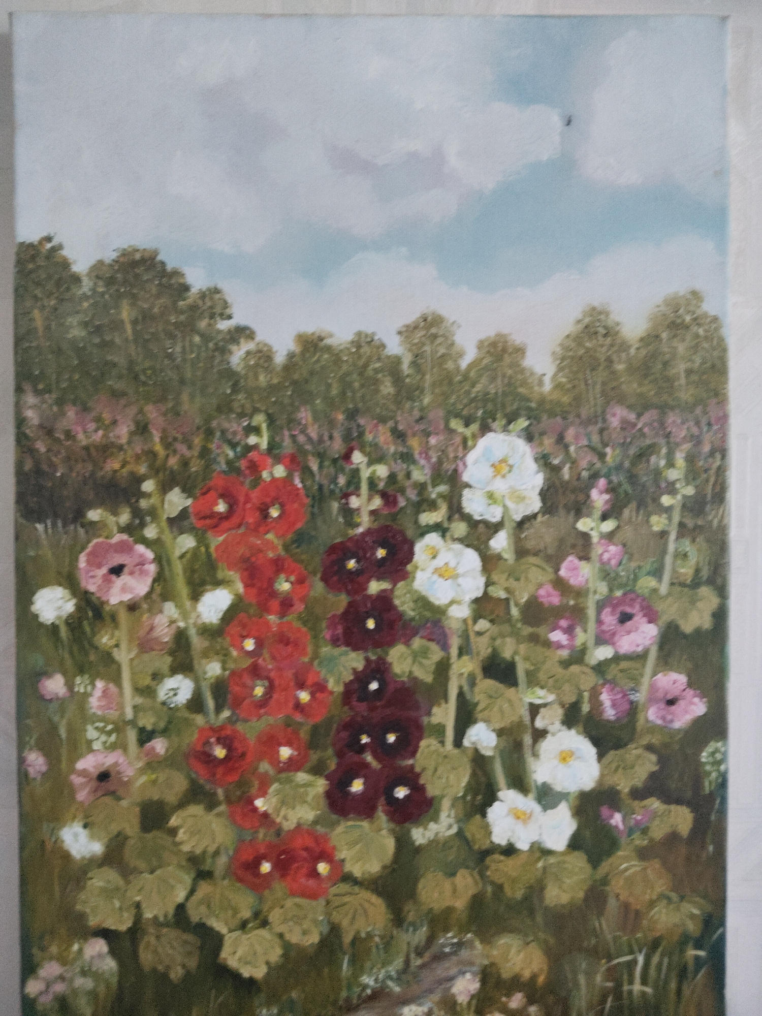A vertical, eye-level, full shot painting depicts a field of hollyhock flowers under a cloudy sky. The flowers, rendered in shades of deep red, burgundy, pink, and white, are the primary focus, with their tall stalks reaching upwards. Behind the hollyhocks, a dense line of trees forms the horizon, their foliage a mixture of greens and browns. The sky above is a pale blue, dotted with soft, white clouds. The overall impression is that of a serene, sunlit summer day in a rural landscape.