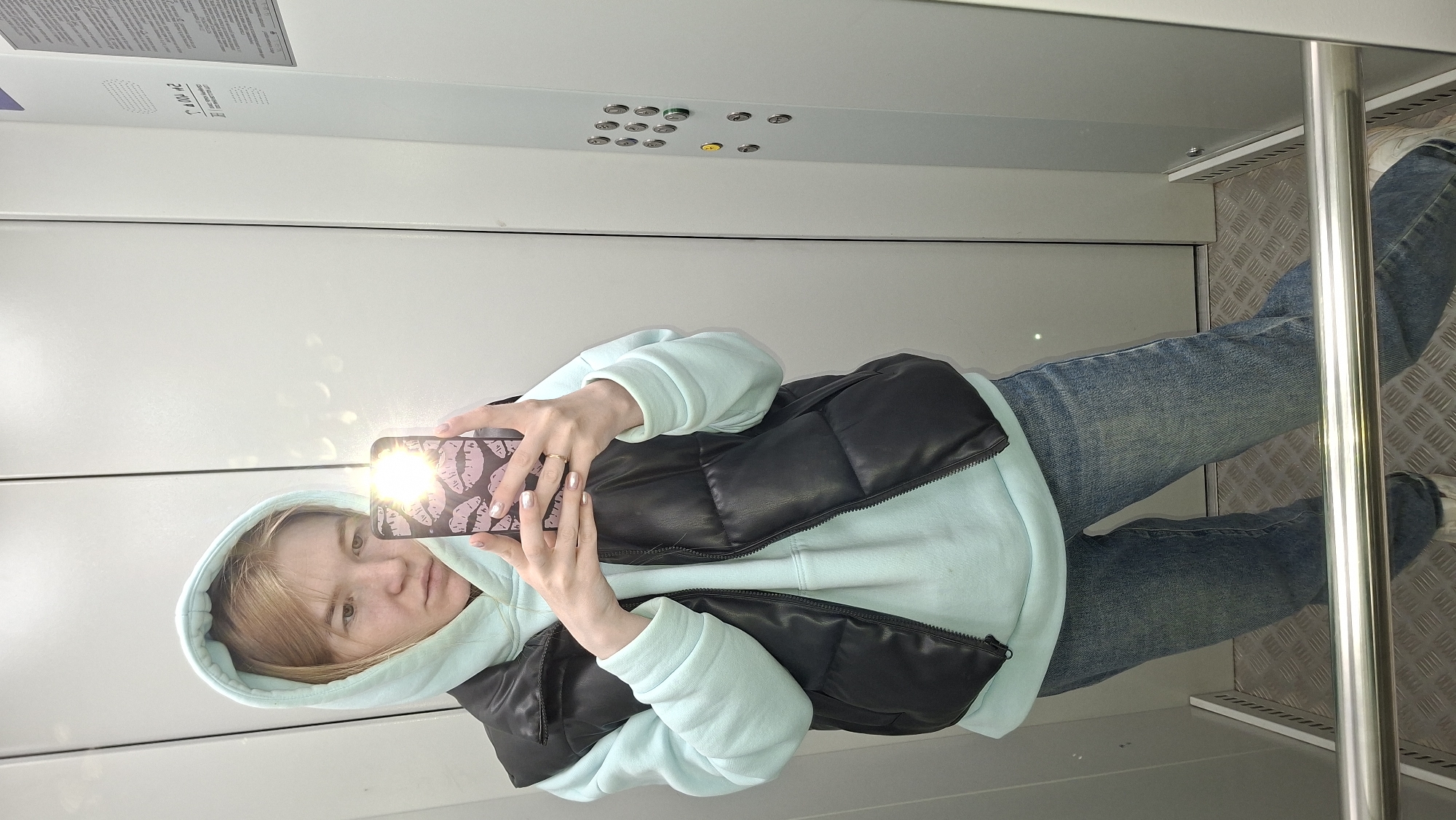 A young woman takes a selfie in an elevator. She wears a light blue hooded sweatshirt, a black puffer vest, and blue jeans. Her phone has a pink case with a lips pattern. The elevator has a panel of buttons and a textured metal floor. The location is Chaikovsky, Russia.