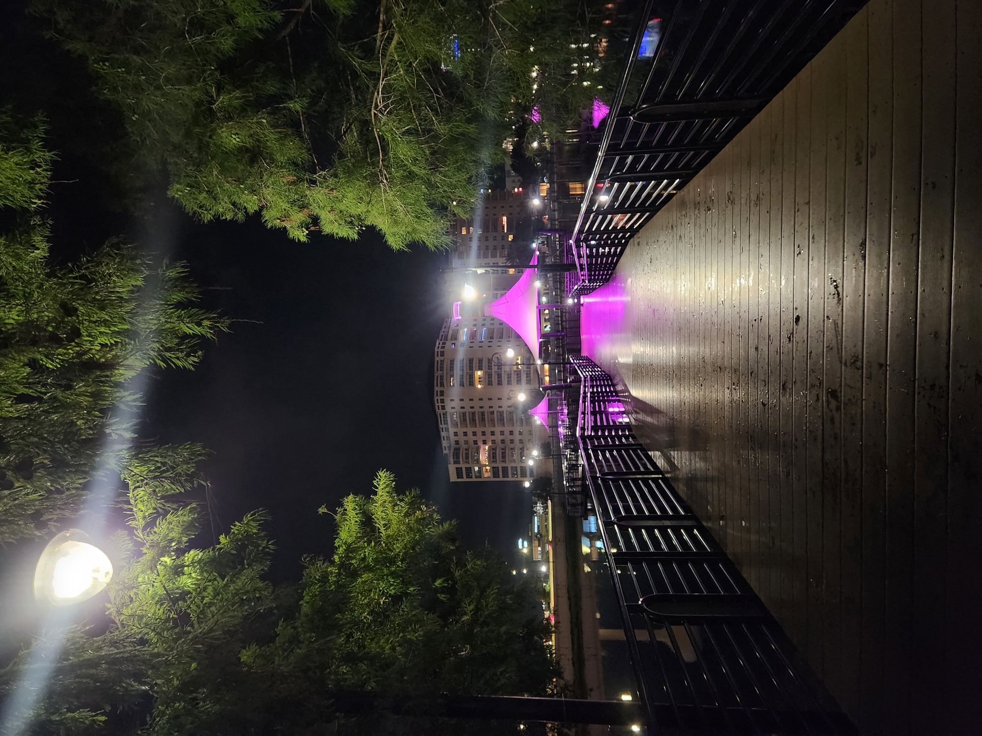 The image depicts a wooden pier at night, illuminated by artificial lights. The pier is wet, indicating recent rain. To the left, a street lamp casts a bright glow through the branches of a large tree. On the right, the pier is bordered by a dark metal railing. In the background, across a body of water, a modern, multi-story building stands with illuminated windows. In front of the building, a vibrant pink tent structure is visible, also lit up. The overall scene is set in a city environment at night, with reflections of lights visible on the water and the wet pier. The location is the Altamonte Springs City Marina in Altamonte Springs, Florida, United States of America. There are no people or animals visible in the image.